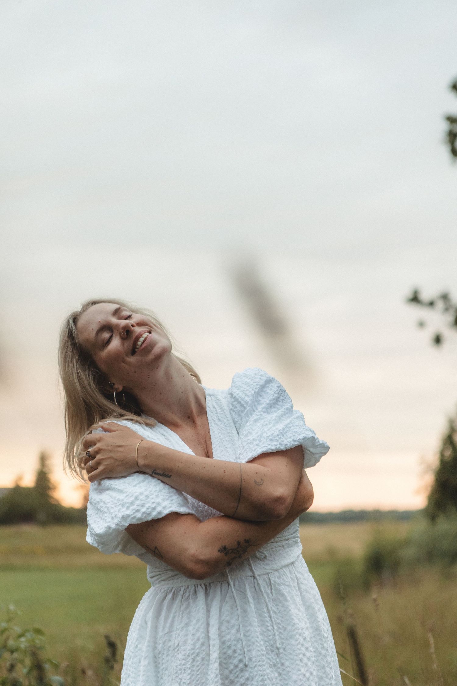 Someone in a white eyelet dress embraces themselves in a golden field at sunset.