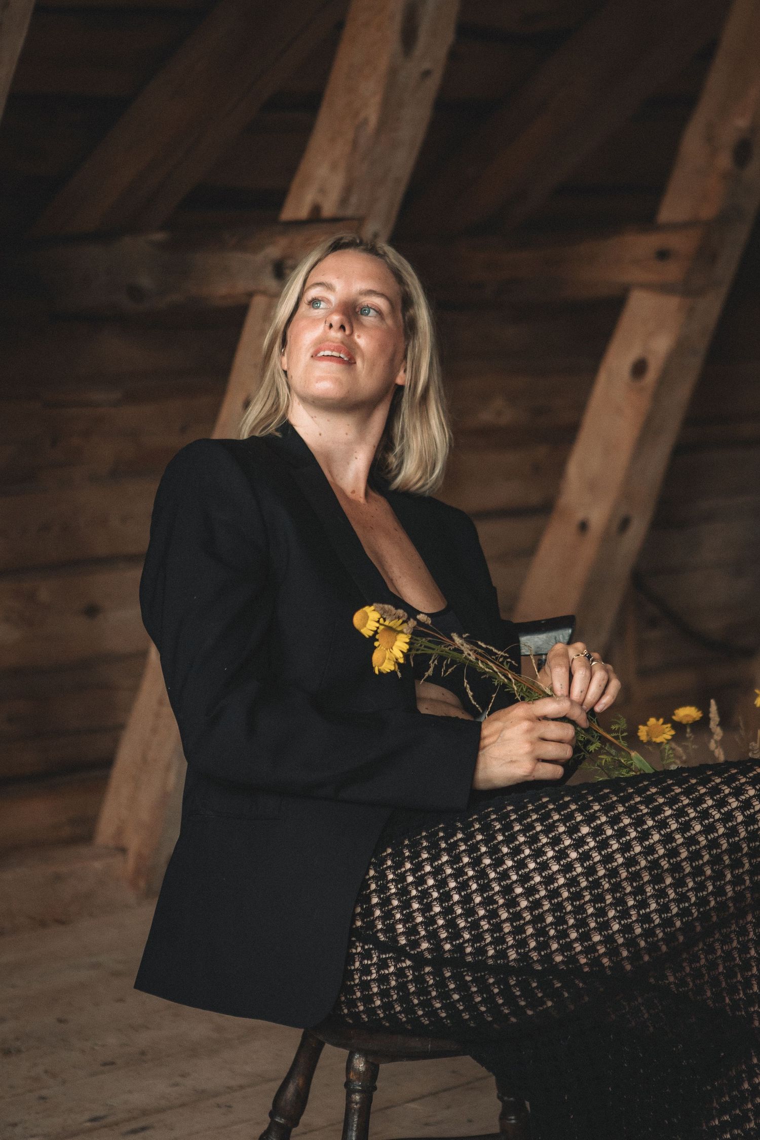 Person in black blazer holding yellow flowers against wooden beam backdrop.
