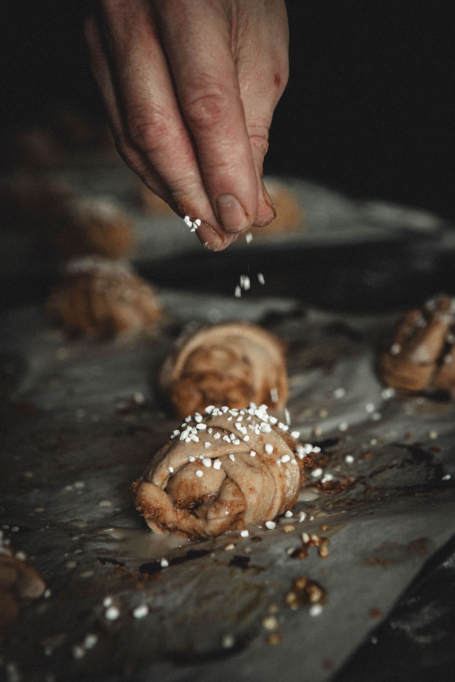 Someone sprinkling pearl sugar onto freshly baked chocolate pastries on a dark surface.