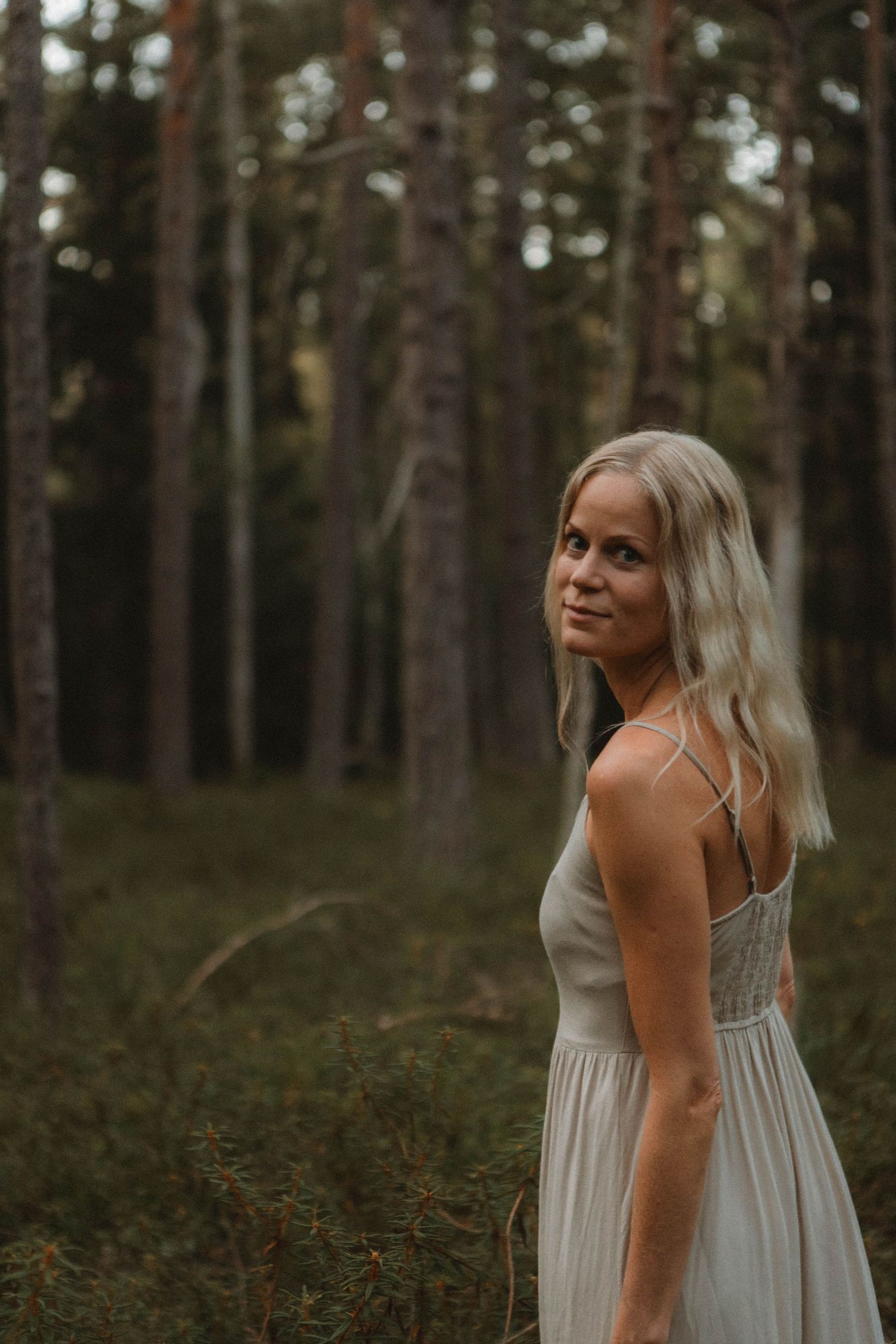 Person in gray dress poses thoughtfully in moody forest setting.