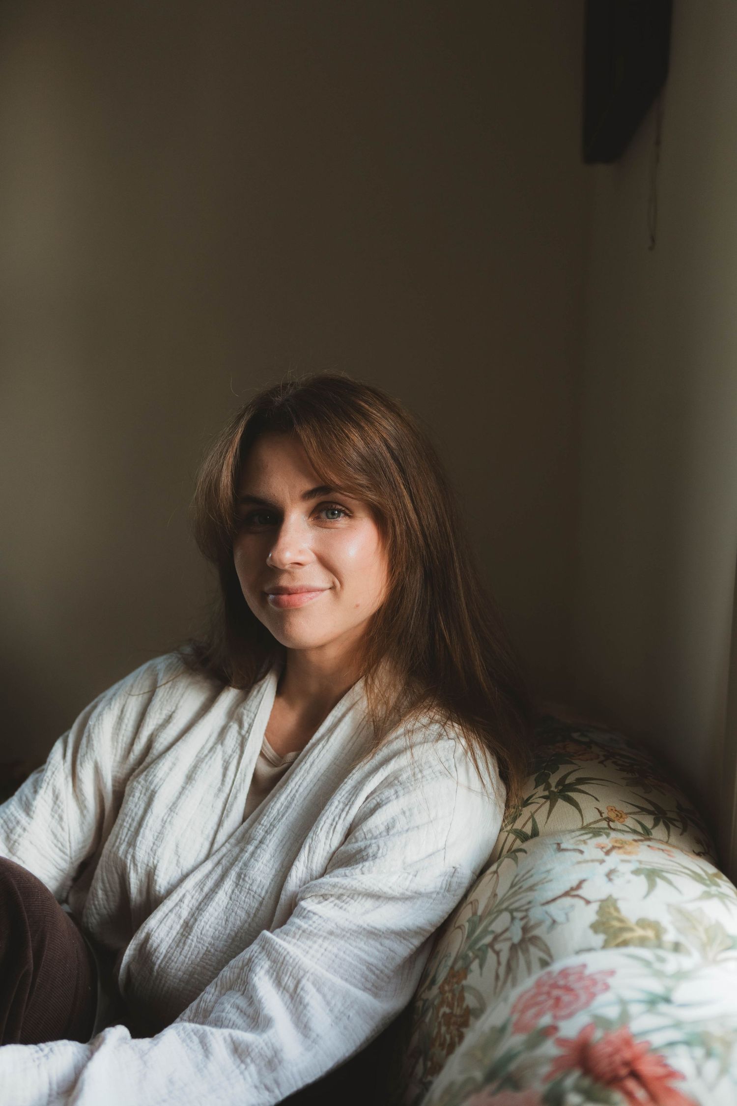 Someone in a cozy white bathrobe relaxes on a floral patterned couch in soft natural lighting.