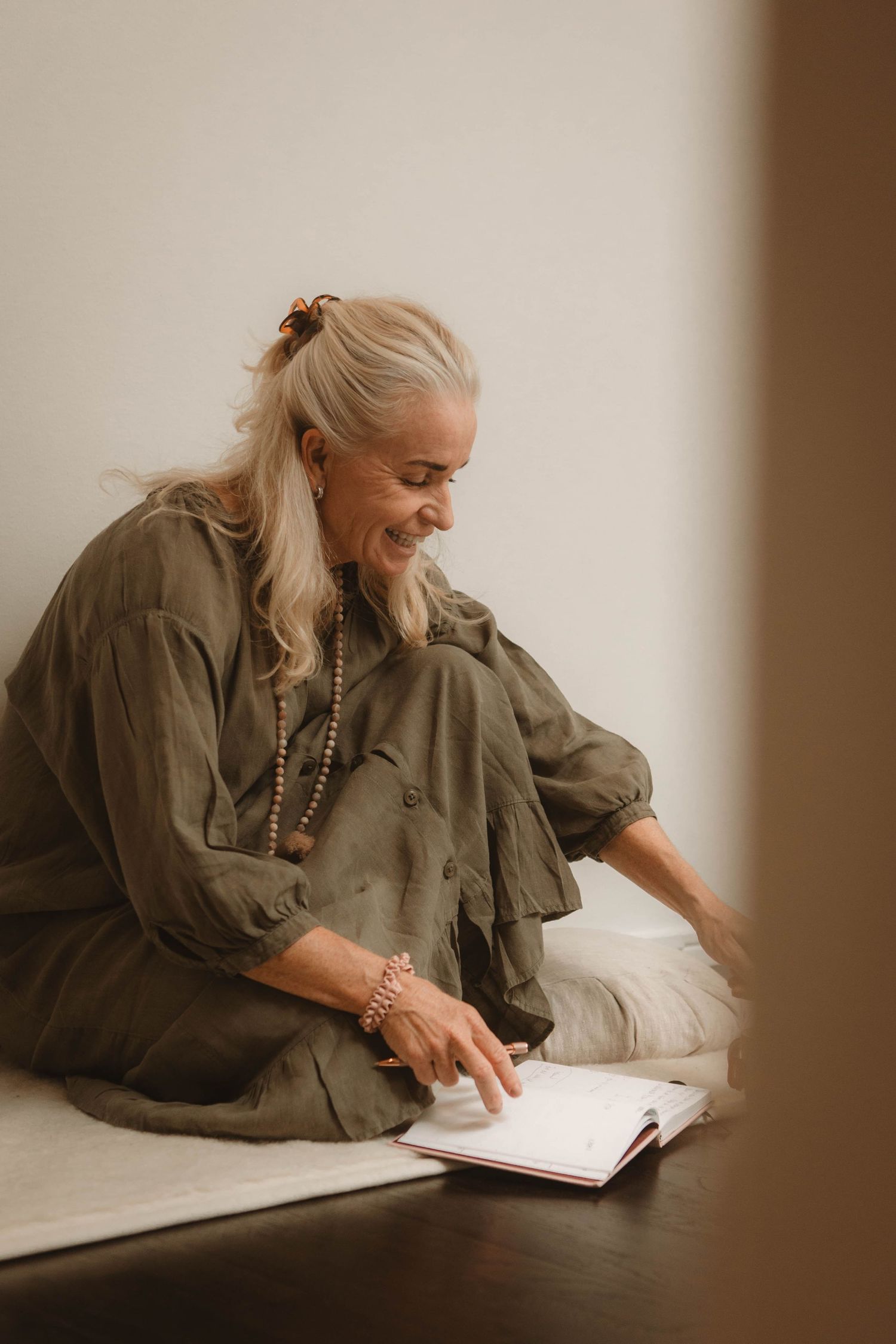 Woman with blonde hair in olive jacket smiling while reading a book in warm lighting.