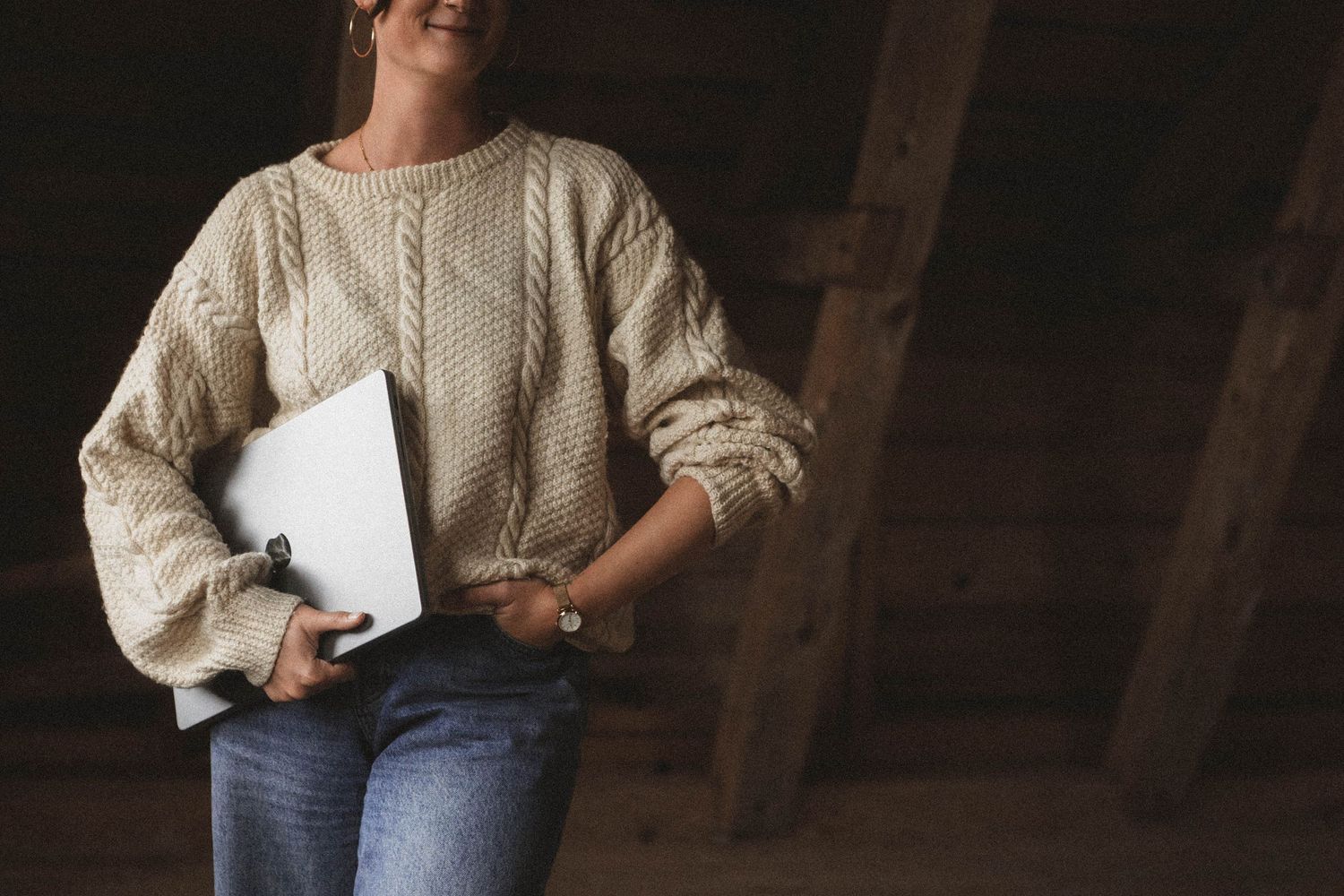 Person wearing beige cable knit sweater and blue jeans poses in indoor setting with dark wood background.