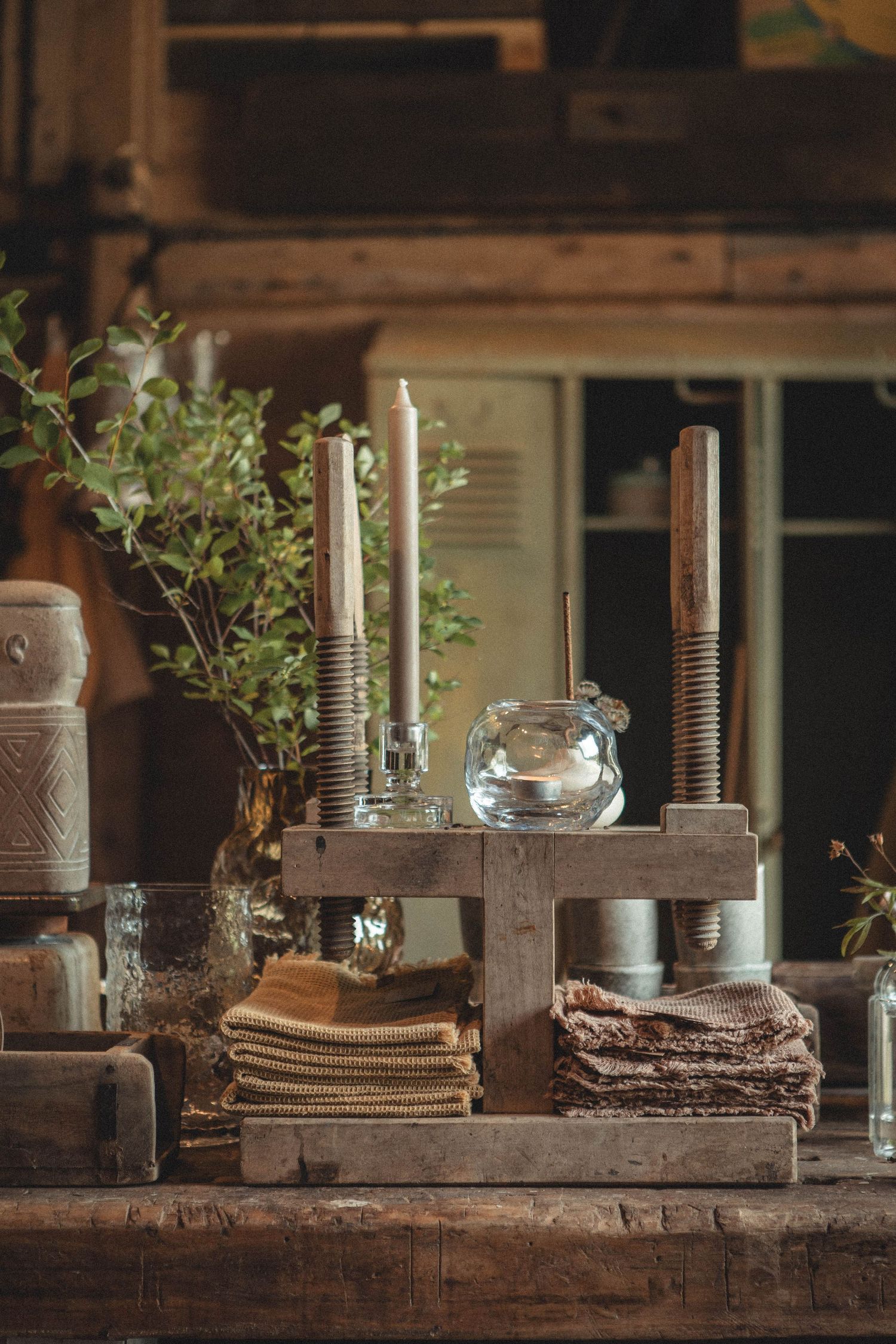 Vintage display with candles, glass containers and folded textiles on wooden table.