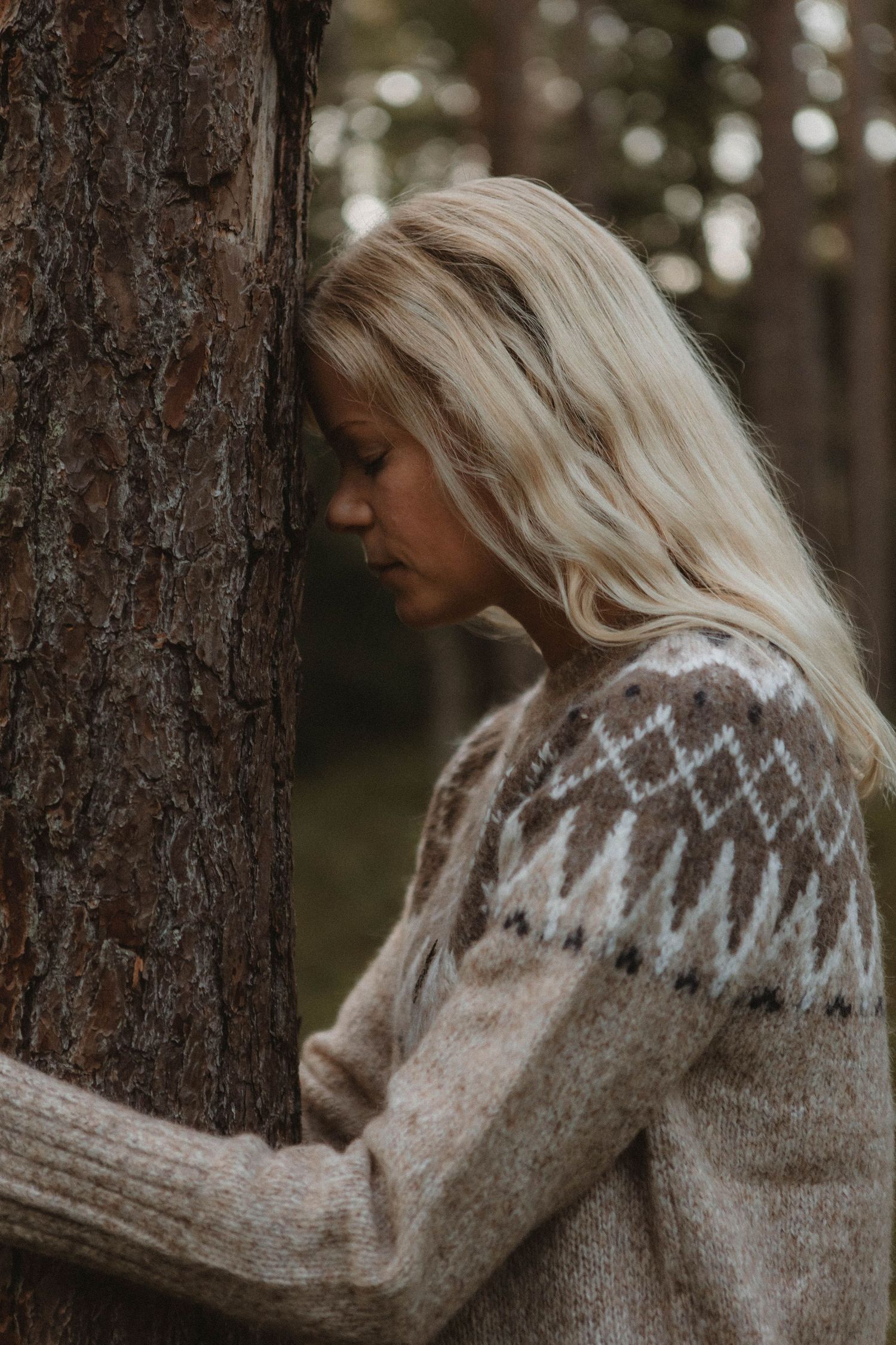 Person wearing a cozy Nordic sweater leans against a pine tree trunk in a forest setting.