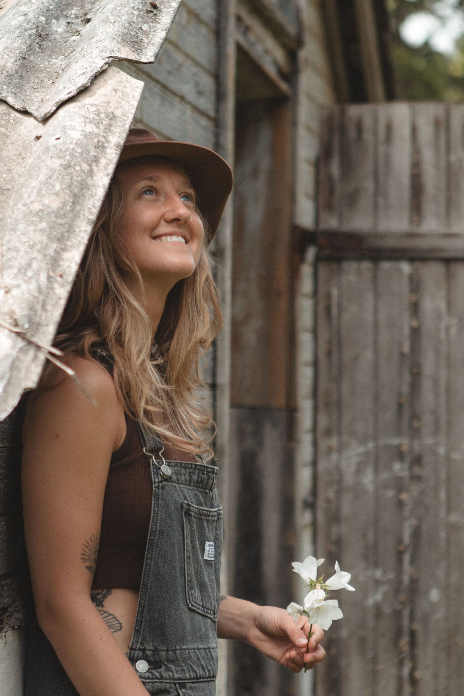 Person in overalls and hat holding white flowers near rustic wooden building.