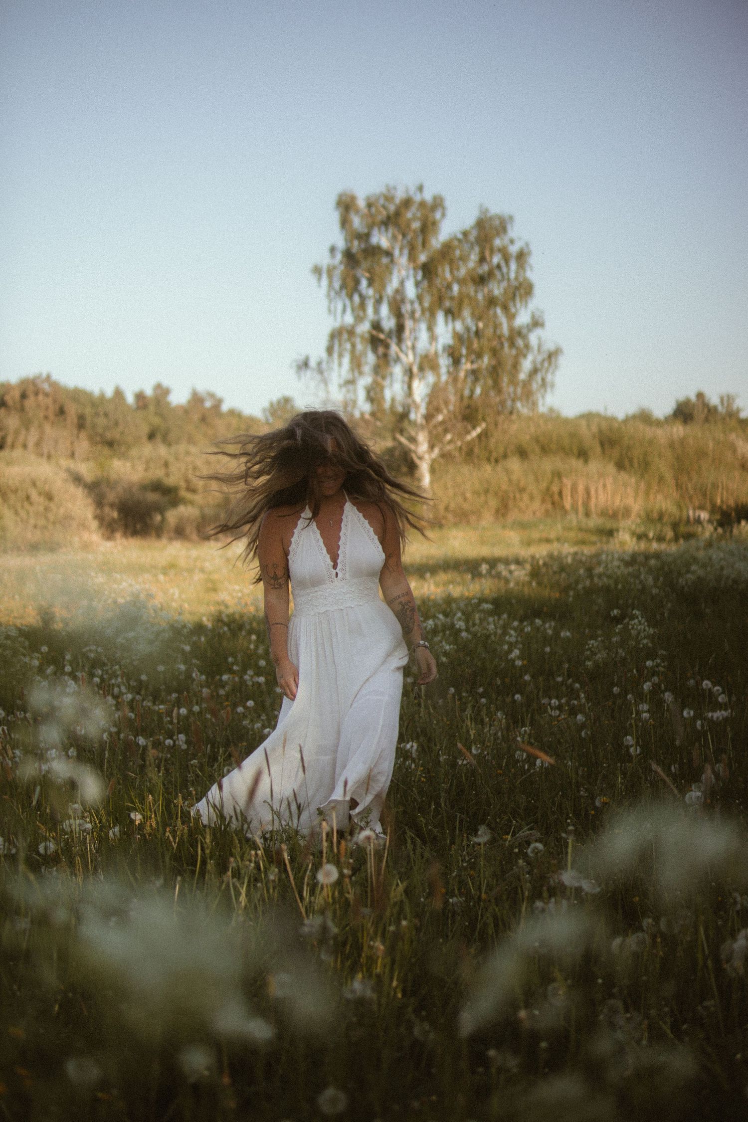Woman in flowing white dress walks through a dreamy summer meadow at golden hour with a birch tree in background.