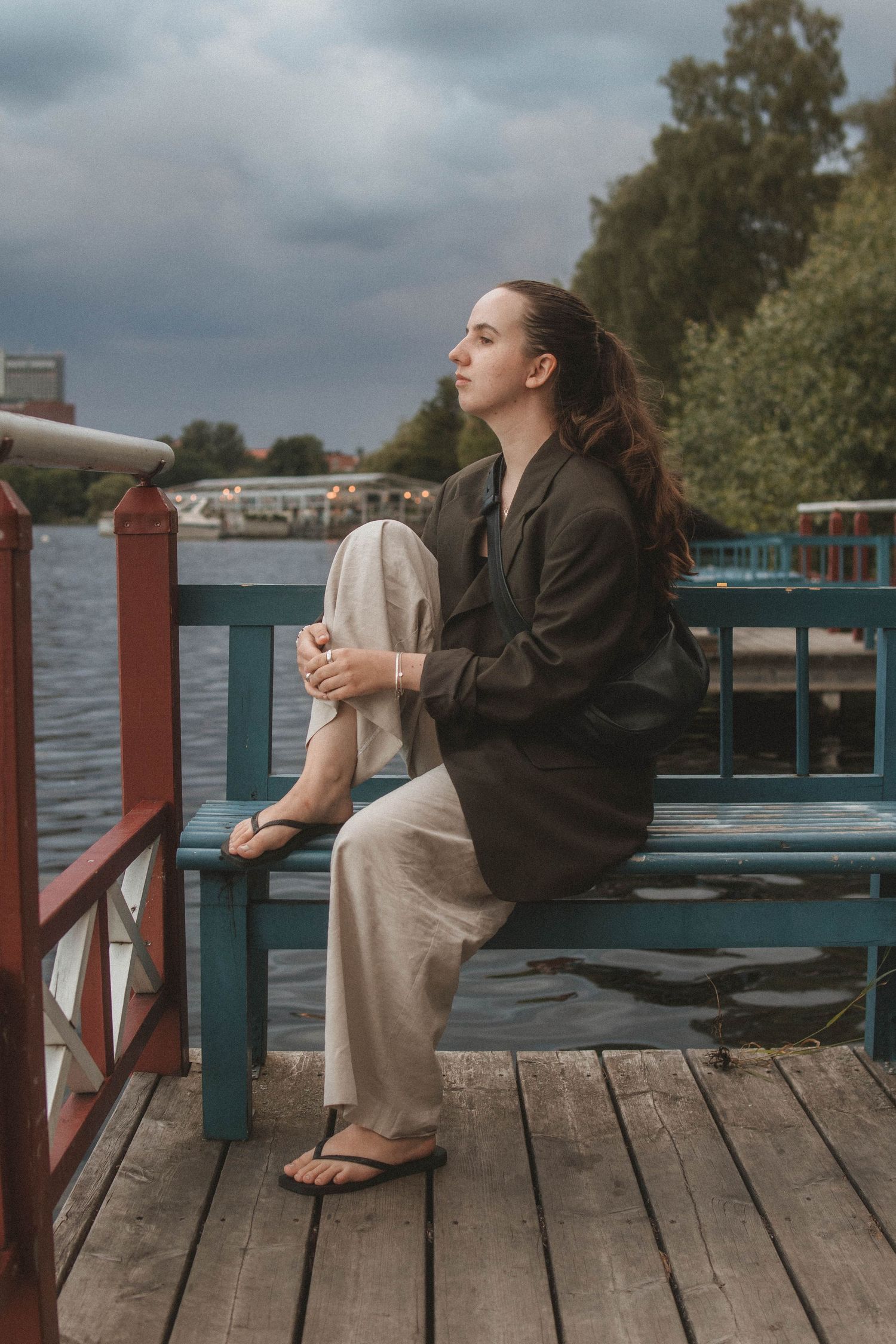 Person in dark jacket and beige pants sits contemplatively on wooden dock overlooking river on cloudy day.