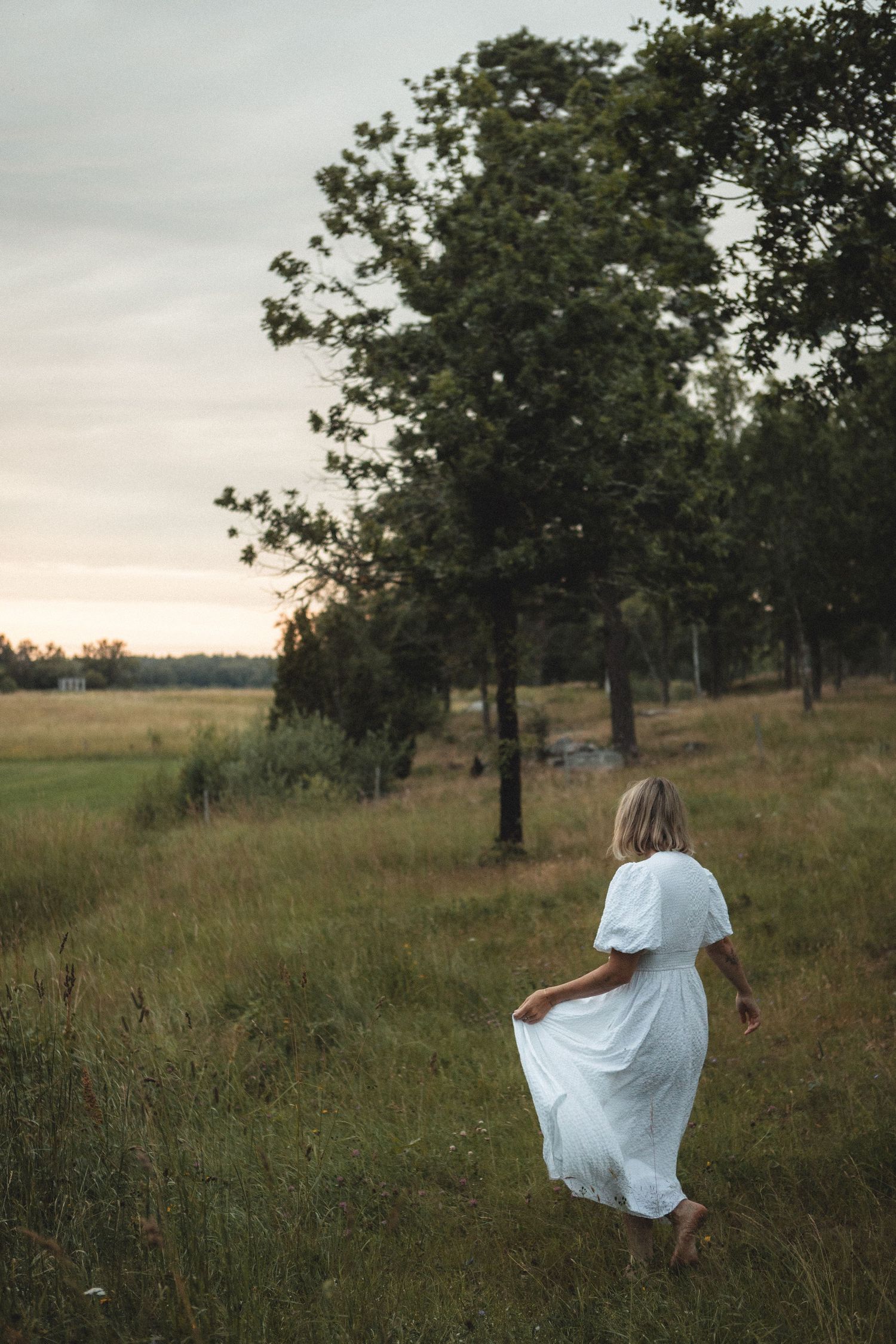 A white dress flows in the breeze as someone walks through a grassy field at sunset.
