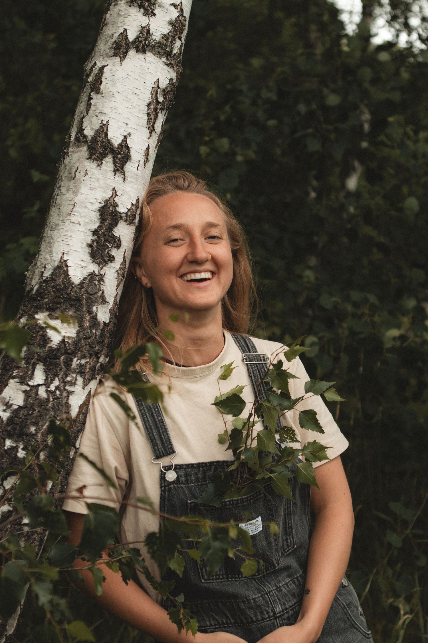 Person in denim overalls smiling joyfully next to a birch tree trunk.
