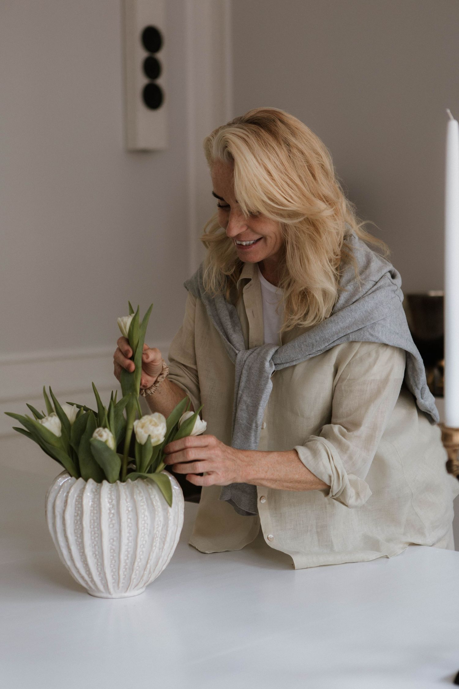 Person arranging white tulips in a decorative ribbed vase on a white table.