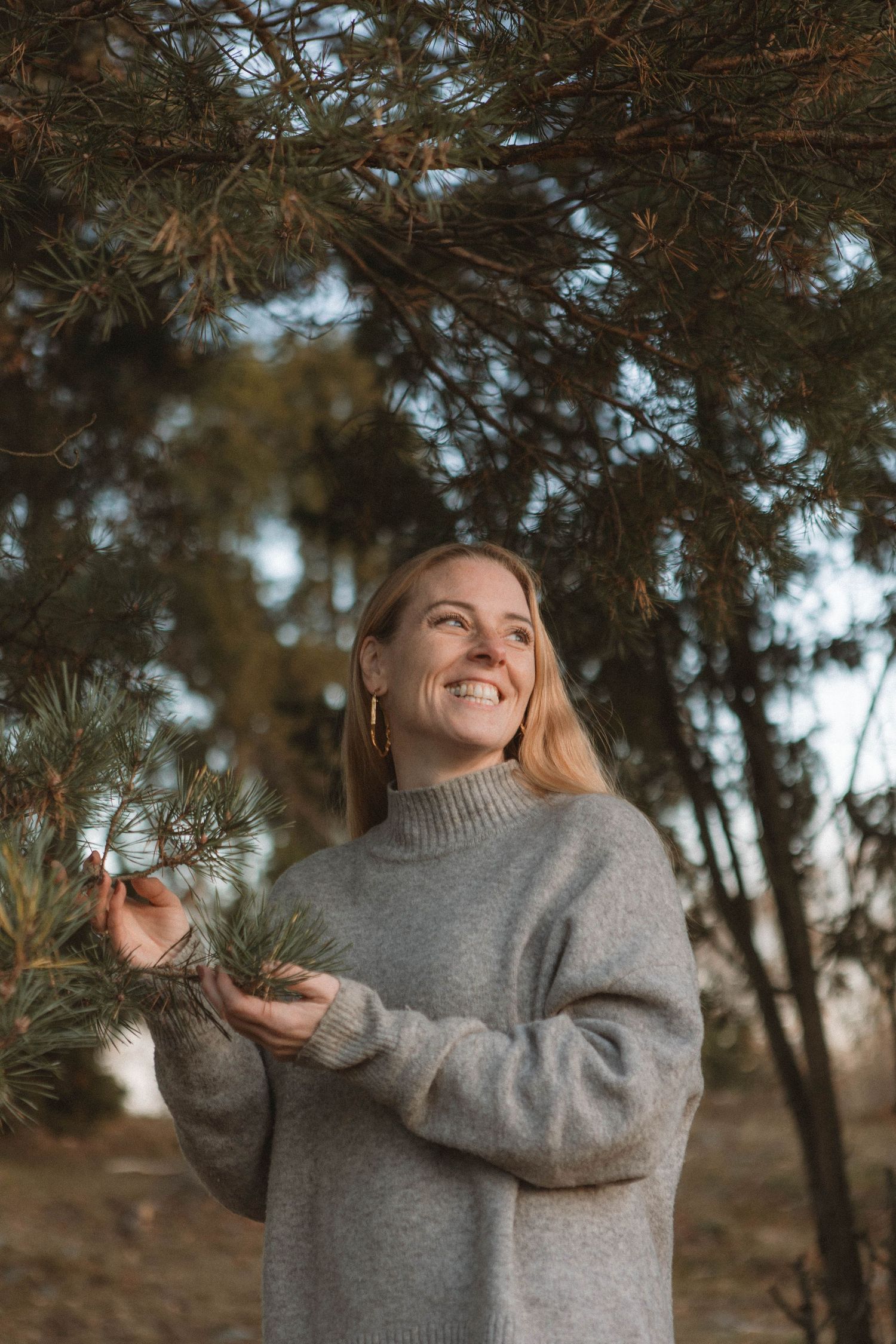 Someone in a cozy gray sweater smiles while touching pine branches on a fall day in the woods.