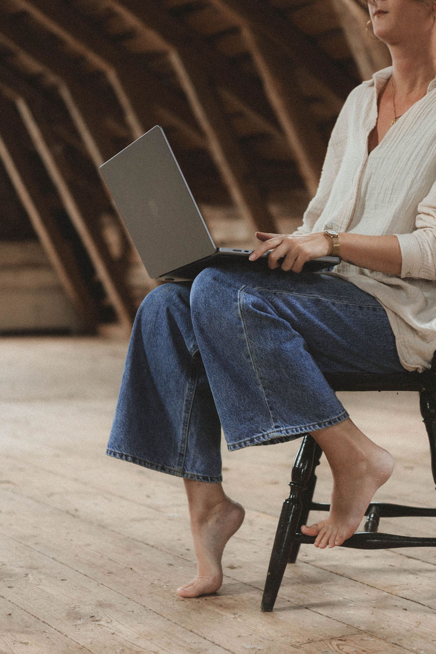 Close-up view of bare feet in blue jeans sitting on a black chair against a wooden attic background.