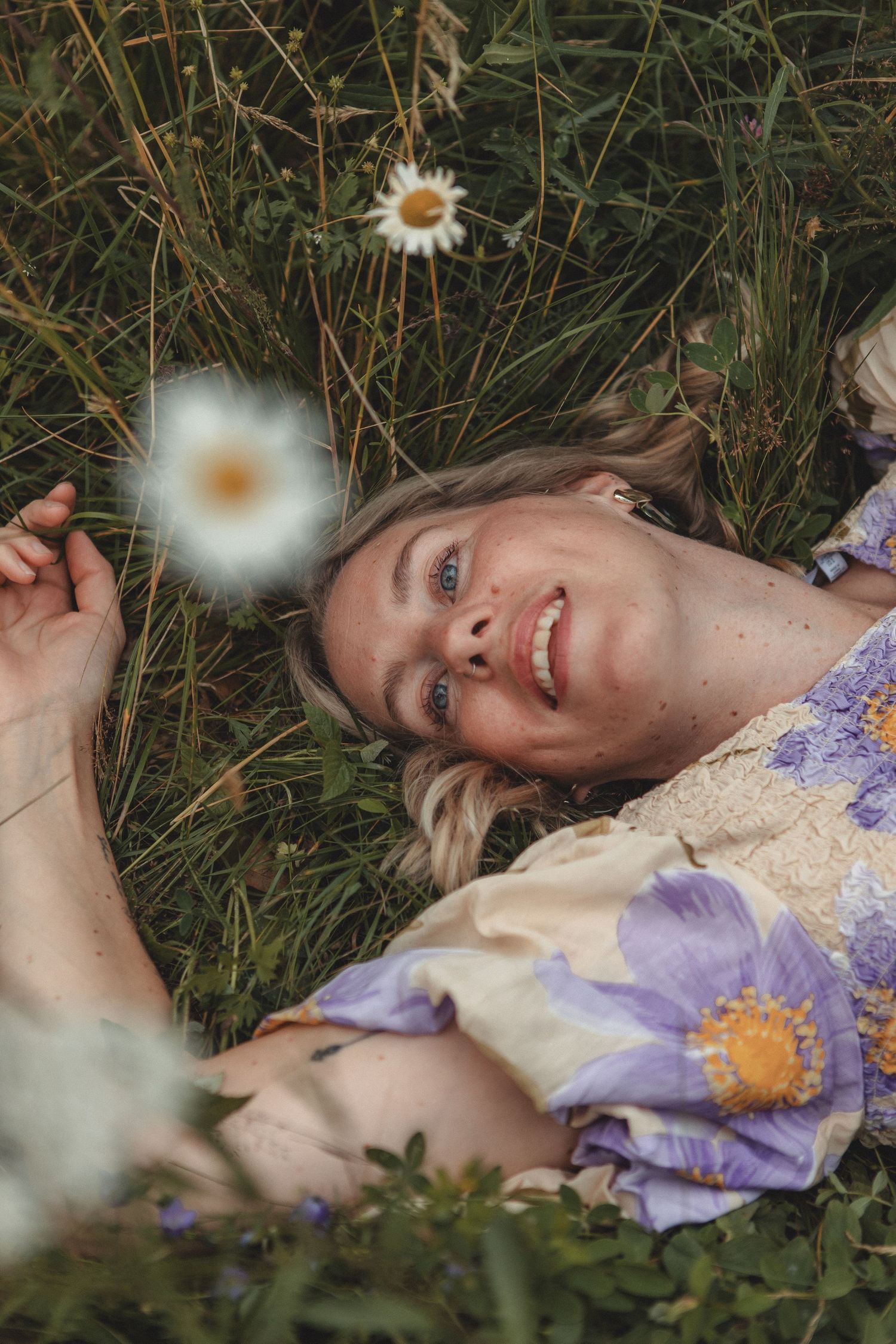 Someone lies in grass surrounded by daisies and purple wildflowers, smiling at the camera.