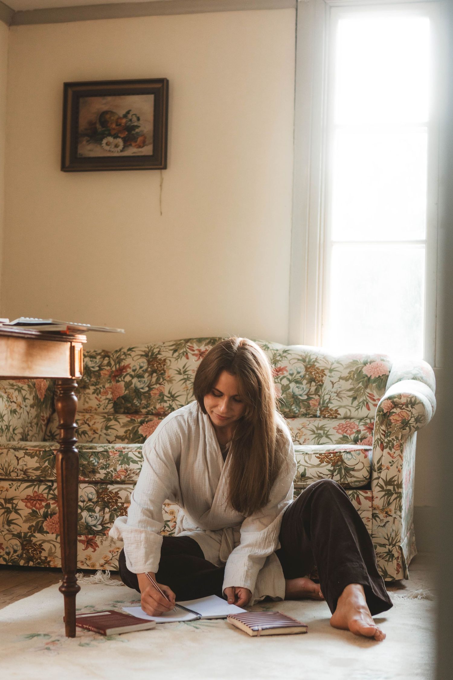 Woman in white sweater lies on floor writing in journal near floral couch in minimalist room with natural light.