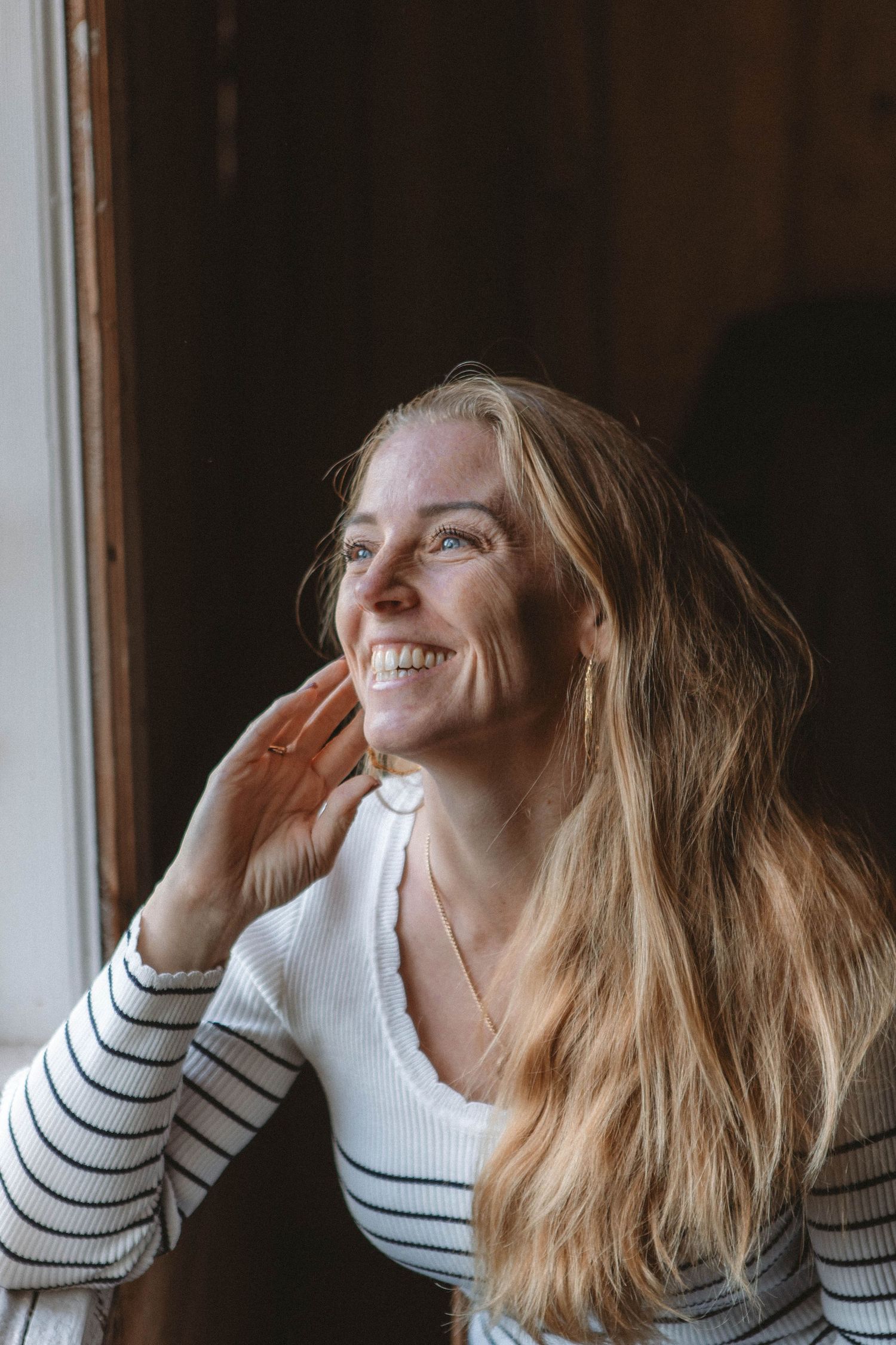 Person wearing striped sweater smiles while looking out window in natural lighting.