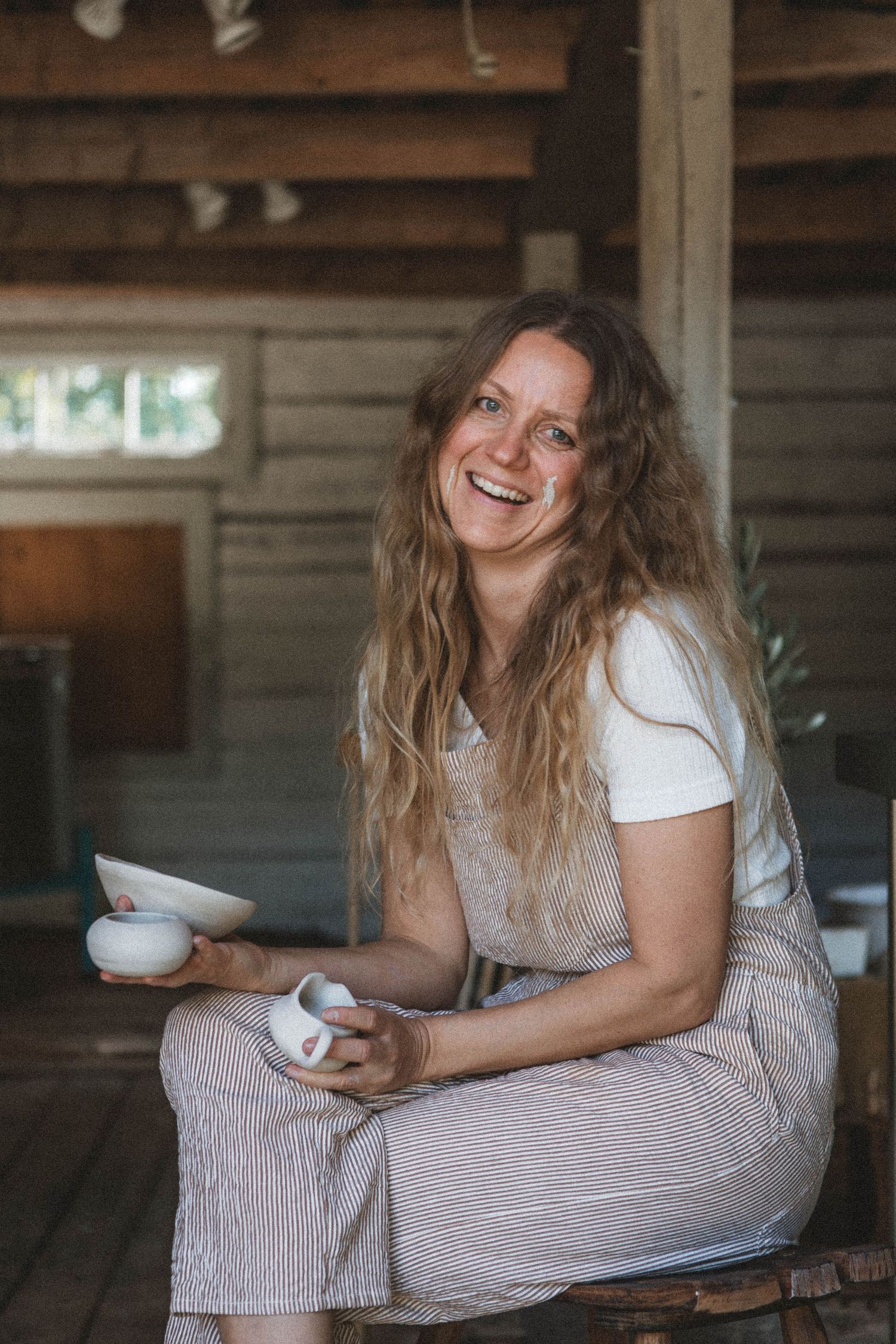 Artist displays handmade pottery while sitting in rustic wooden studio space.