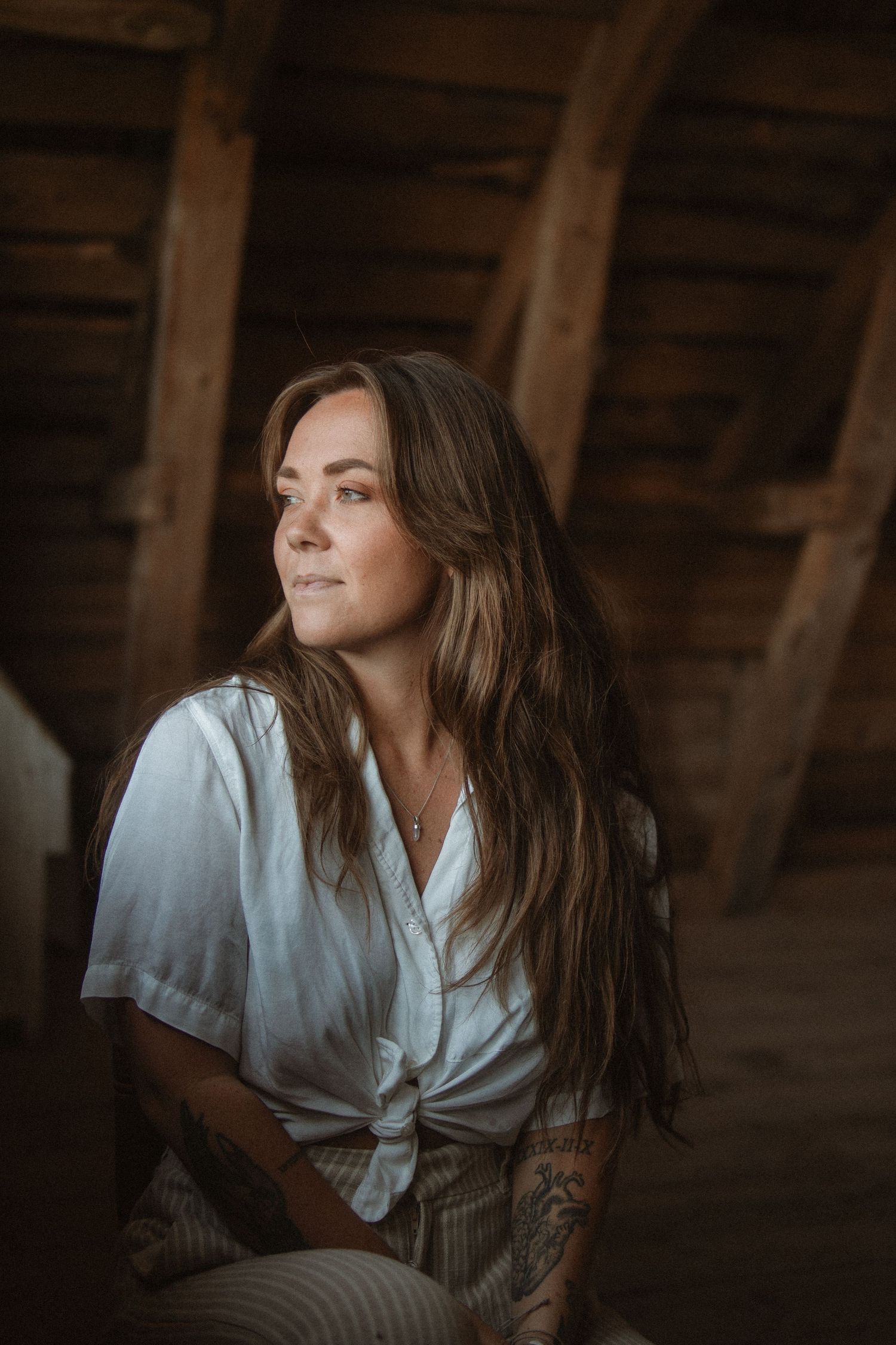A person with long brown hair sits in a rustic attic wearing a white tied shirt looking to the side in moody lighting.