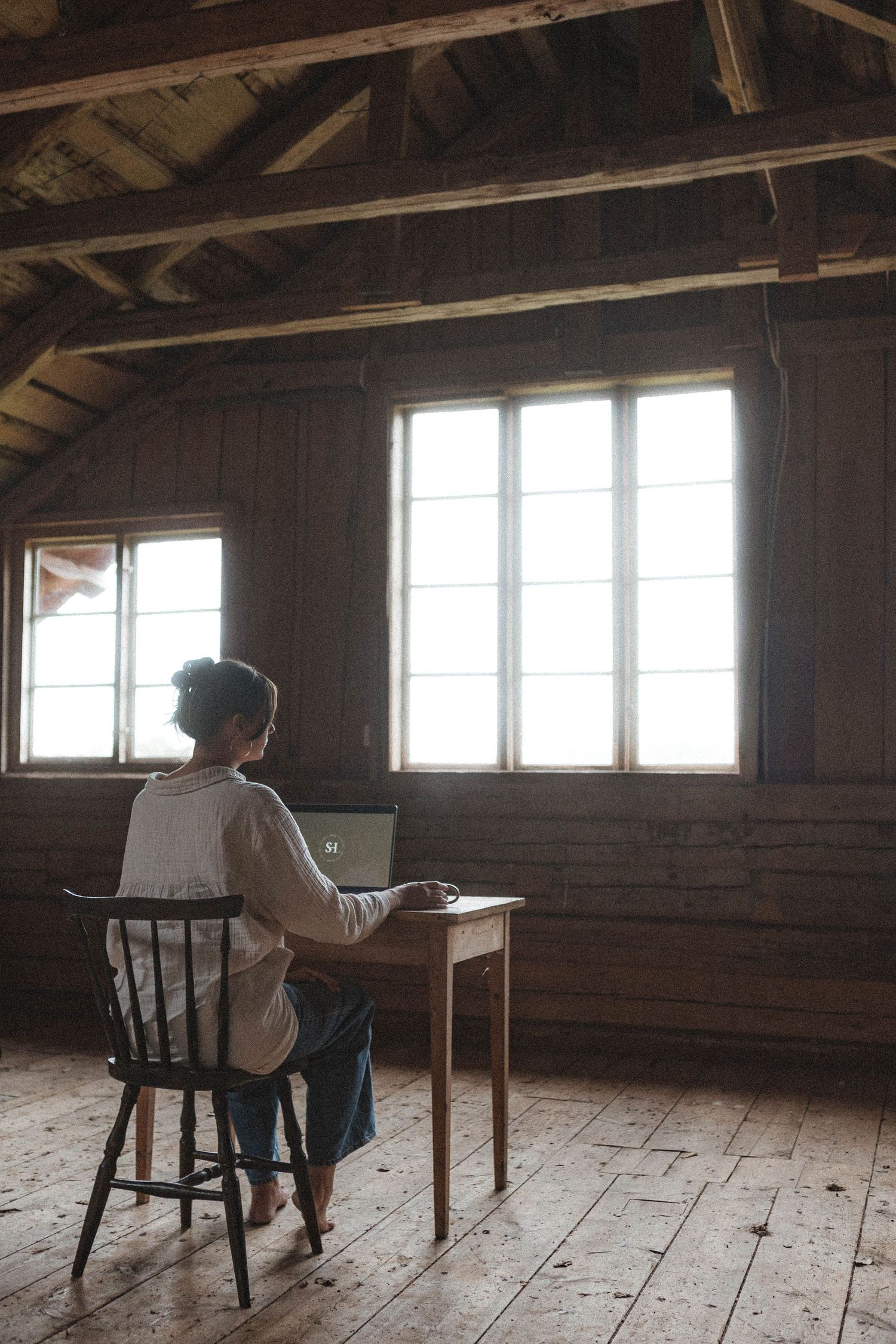 Table and chair silhouetted against bright windows in rustic attic workspace.