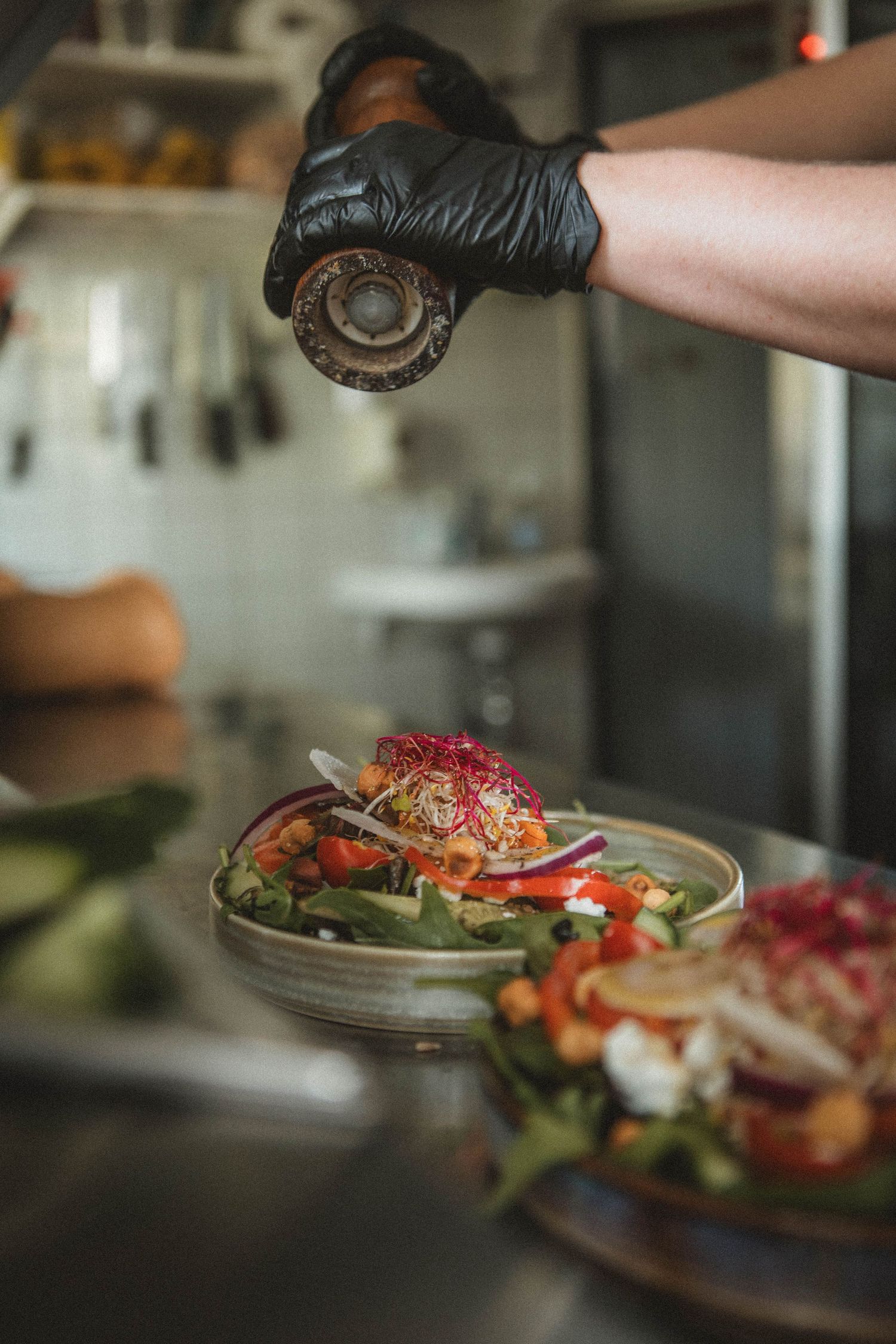 Chef wearing black gloves seasoning a fresh salad with spices in a restaurant kitchen.