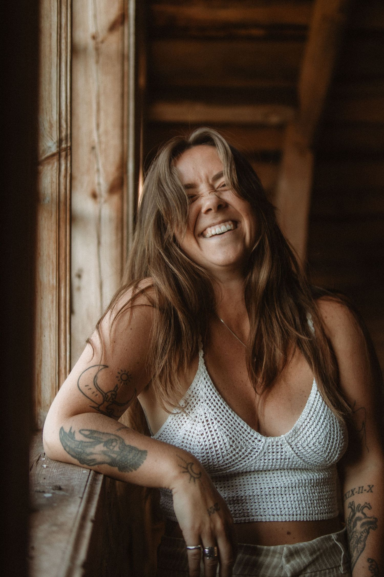 A joyful portrait in warm sepia tones shows someone in a white crochet crop top with tattoos, laughing against wooden walls.