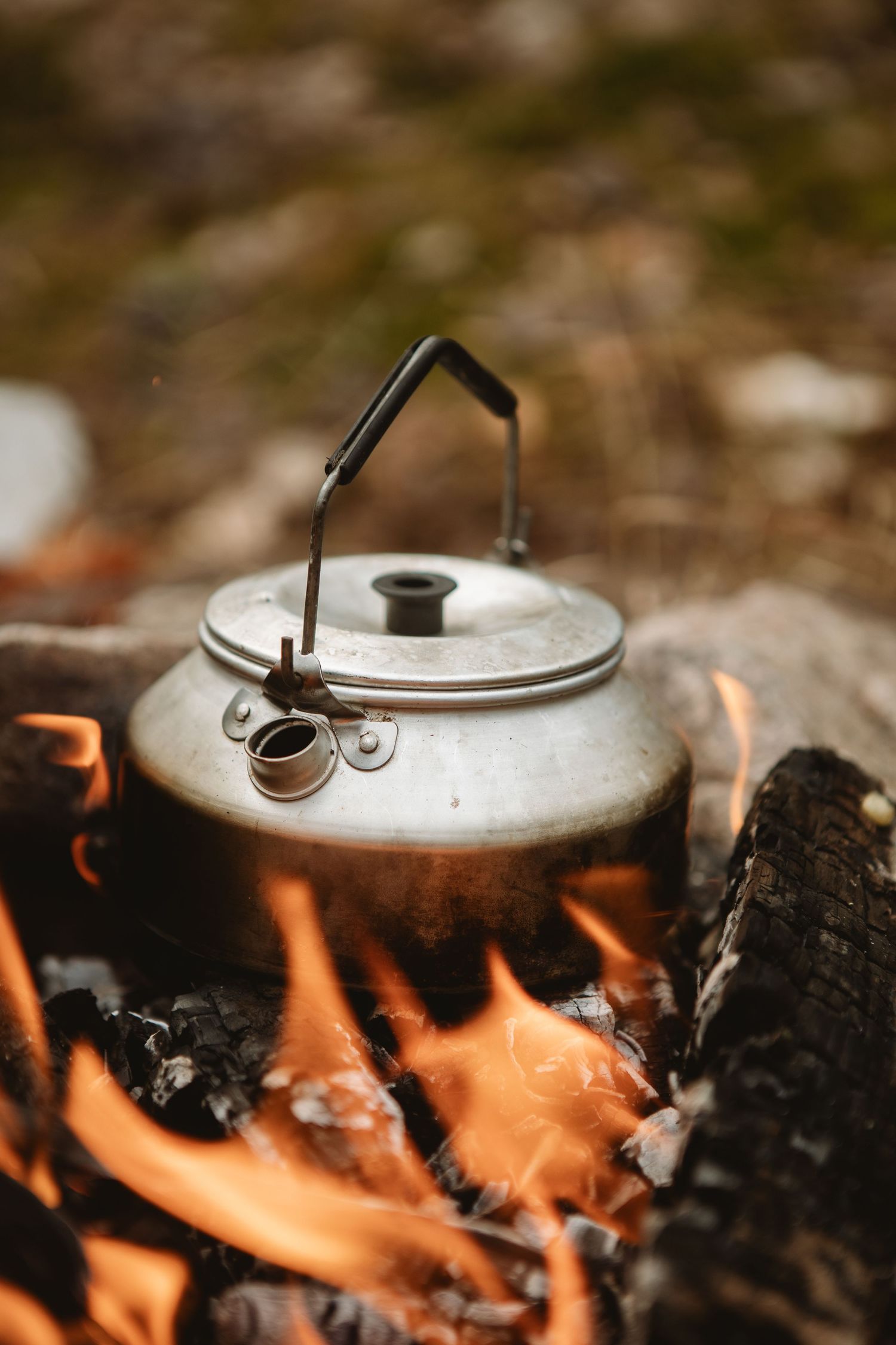Vintage aluminum kettle heating over open campfire flames on burning logs outdoors.