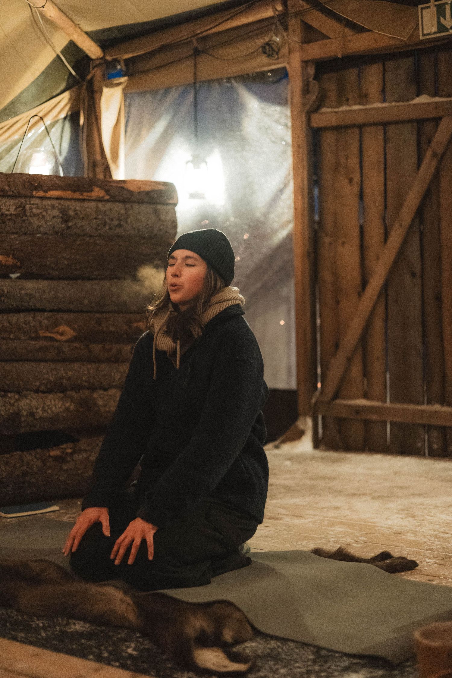 A person sits on a floor mat in a rustic cabin-like structure with wooden walls and beams during a meditative moment.