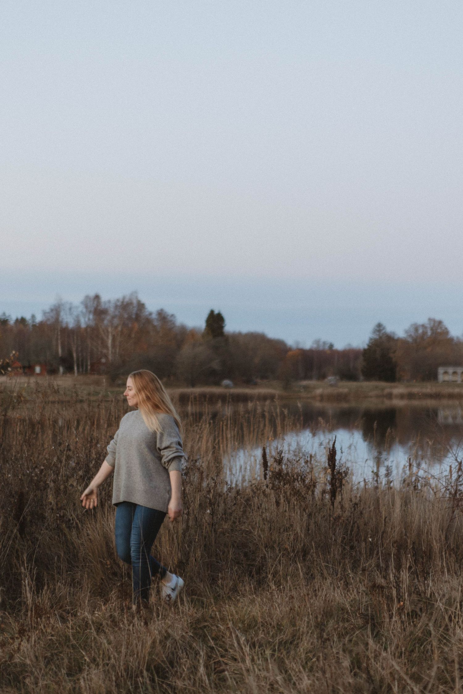A person stands in tall brown grass by a serene lake at dusk, wearing a grey sweater and jeans in an autumn landscape.