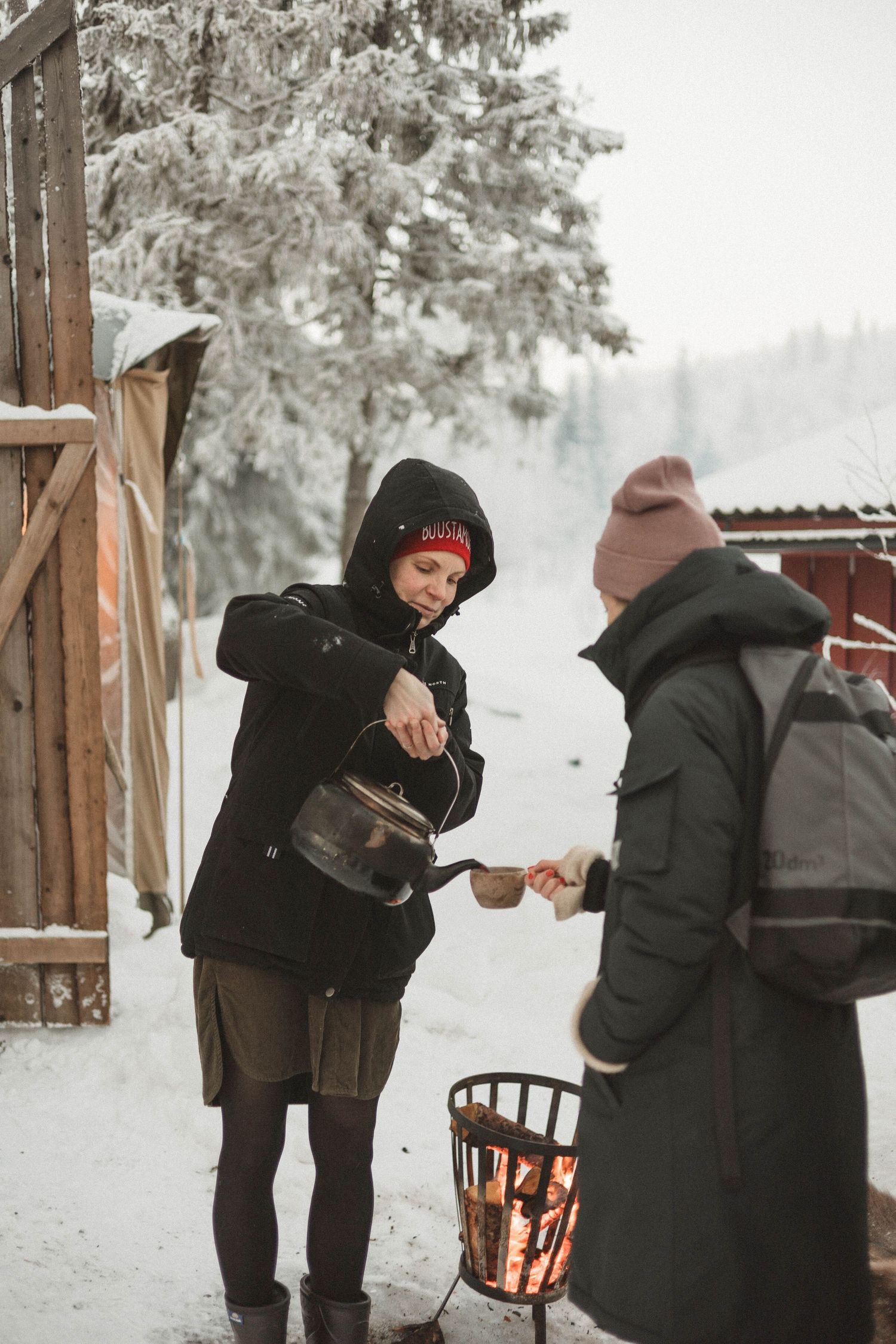 Two people in winter coats and hats stand together in snowy outdoor setting near a fire basket.