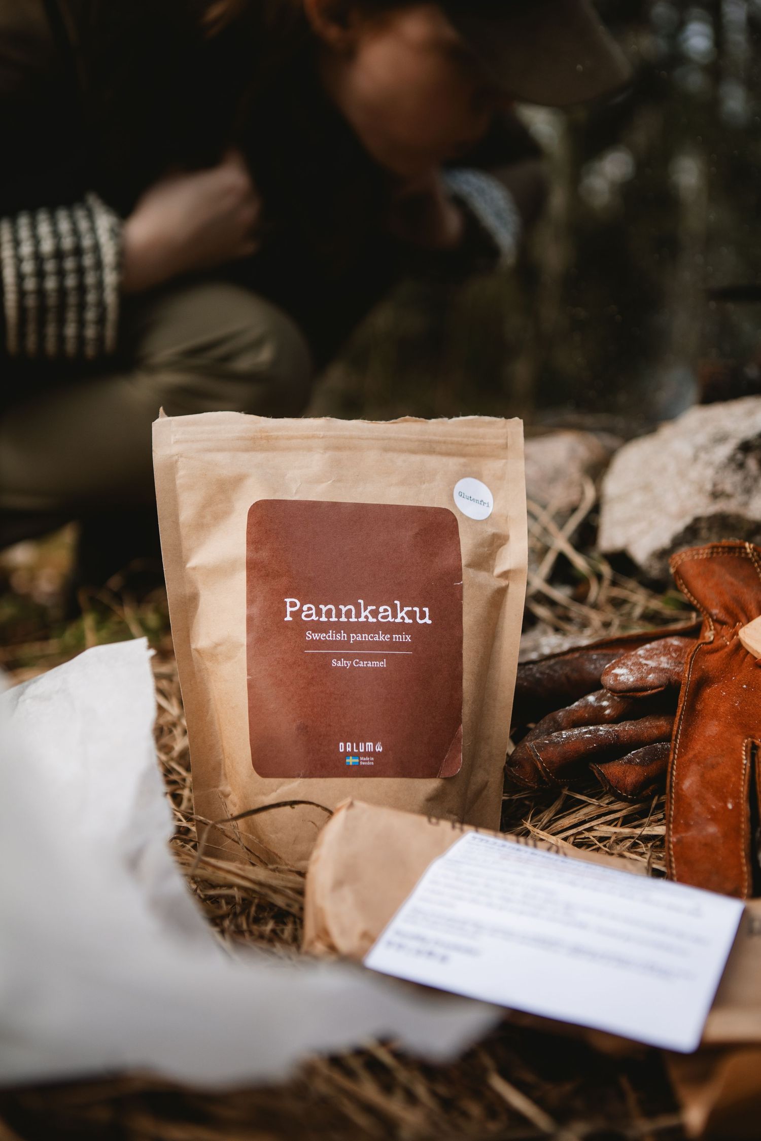 Pannkaku Swedish pancake mix in kraft paper bag displayed outdoors on hay next to sausages in forest setting.