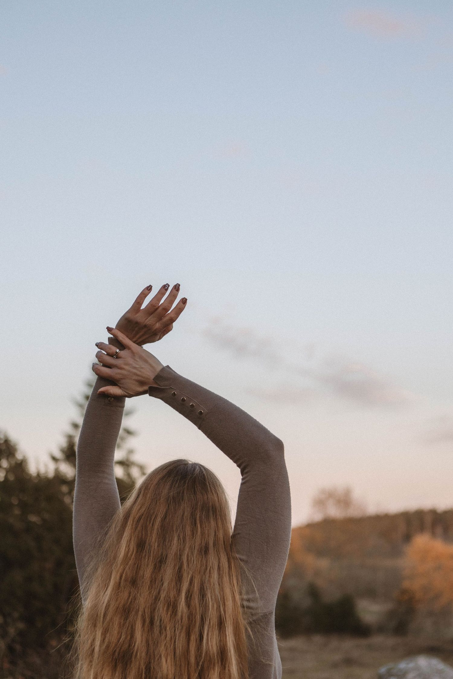 Person with long hair raises arms toward sunset sky in a peaceful nature setting.