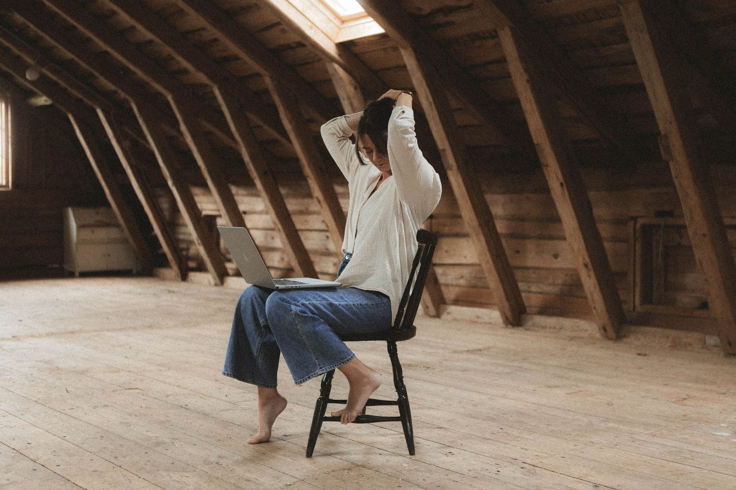 Two wooden chairs sit in an empty rustic attic with exposed wooden beams and natural light coming through a skylight.