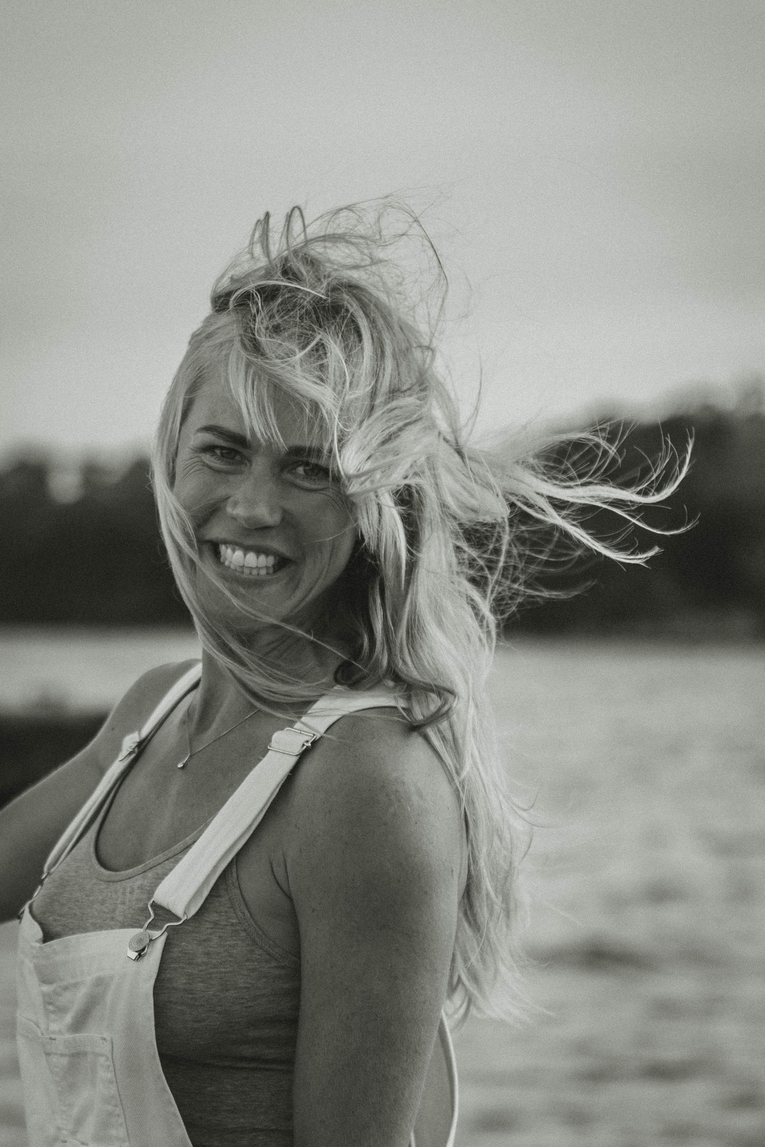 Candid black and white portrait of someone with windswept hair wearing overalls by the water.