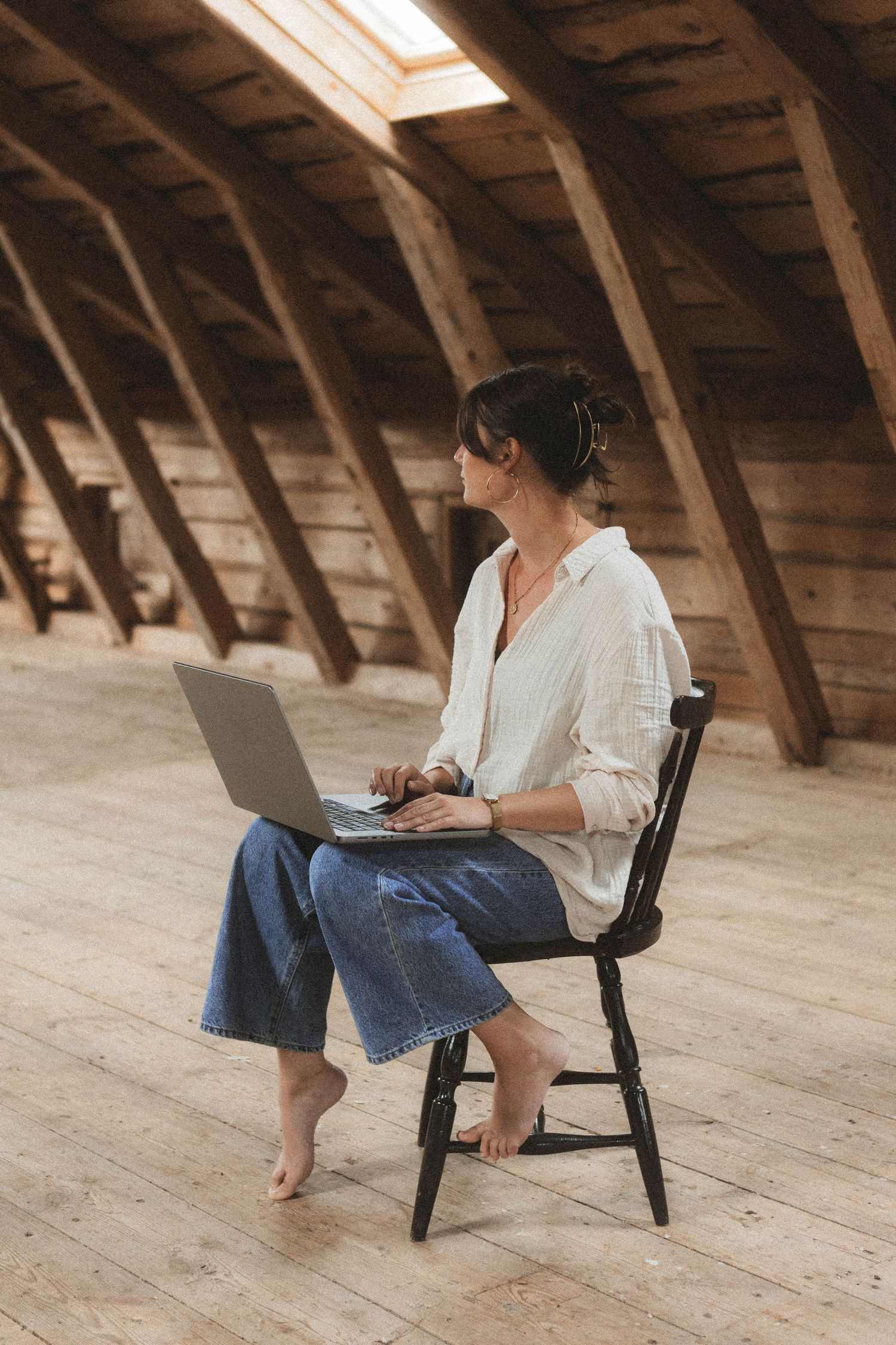 Person sitting in wooden attic space working on laptop in casual attire with exposed wood beams overhead.