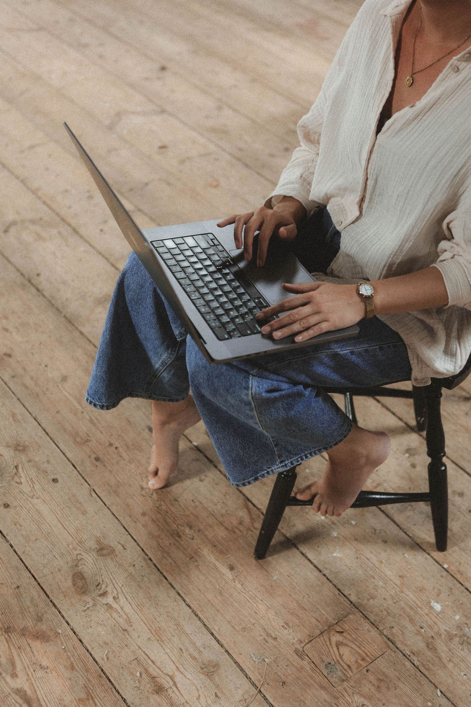 Person wearing blue jeans types on gray laptop computer while sitting on wooden floor with bare feet.