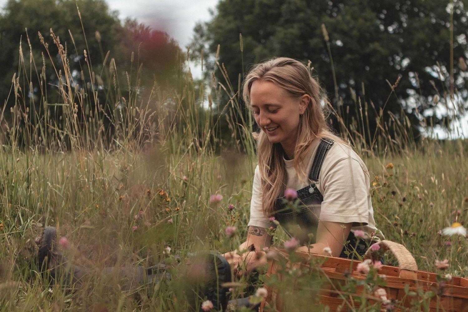 Person gathering flowers in rustic basket among tall grasses and wildflowers.