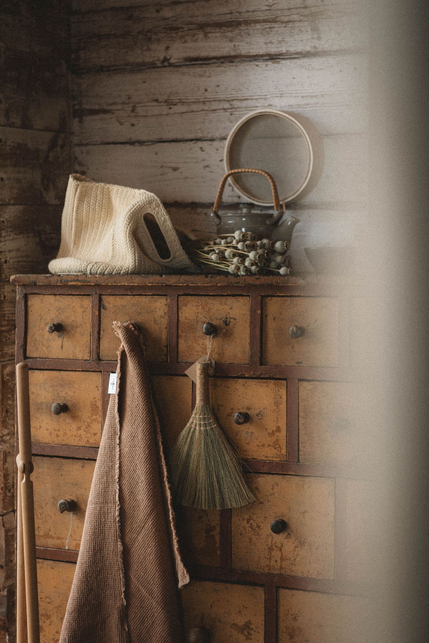 Antique wooden drawer unit with hanging broom and vintage tea kettle in rustic setting.