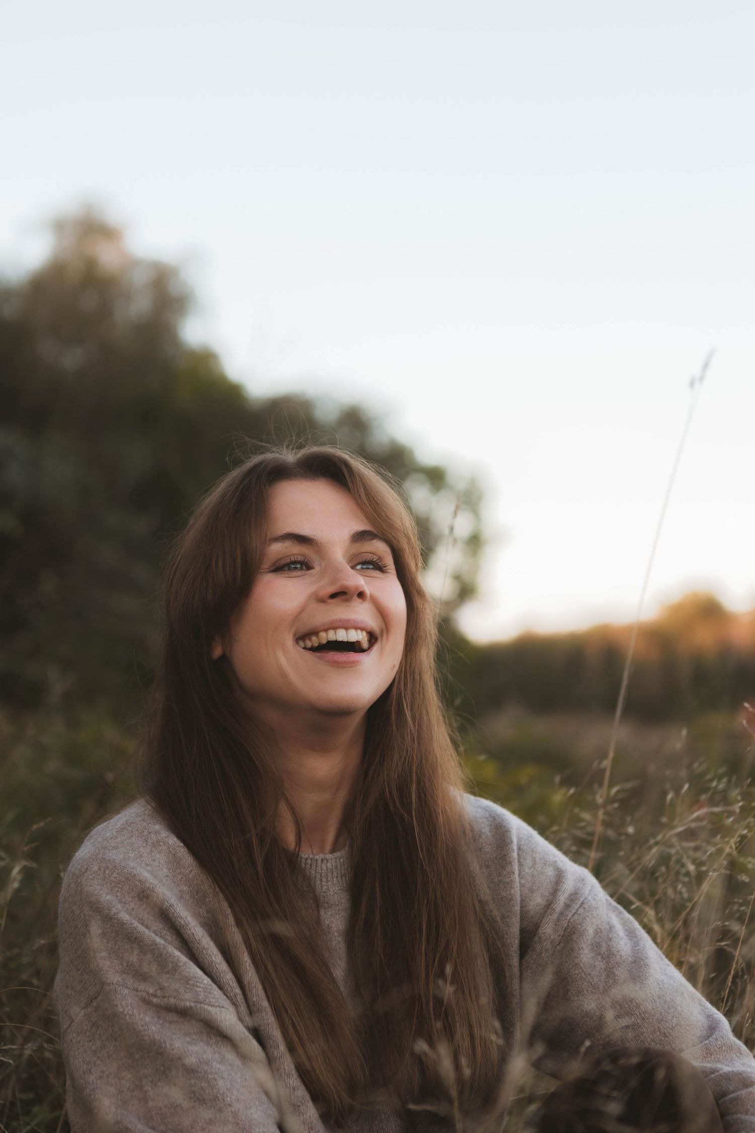 Person laughing joyfully while sitting in a grassy field at sunset wearing a cozy gray sweater.