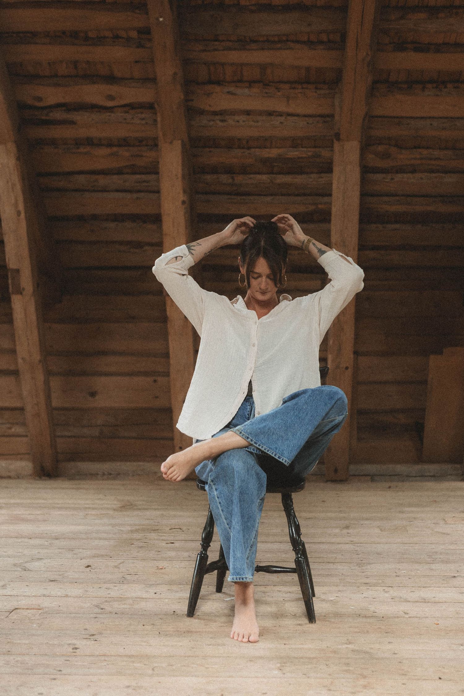 Person in white button-down shirt and jeans sits barefoot on wooden stool under rustic wooden ceiling.