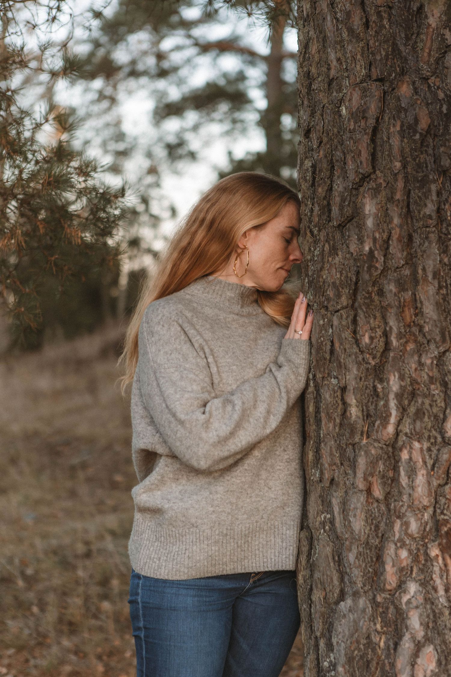 Someone in a gray sweater and jeans leans against a large tree trunk in a moody outdoor autumn setting.