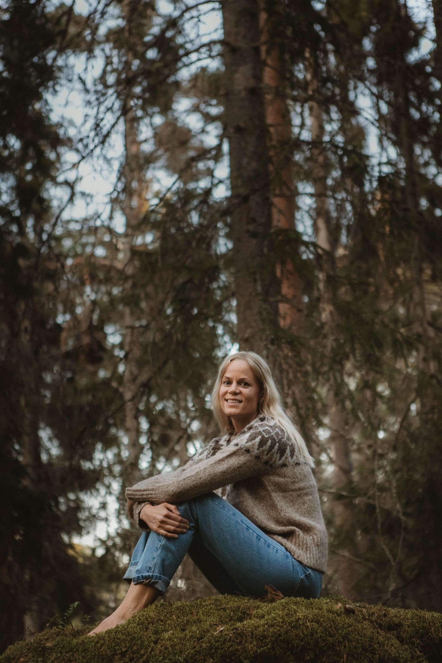 Person in knit sweater and jeans sits peacefully on mossy ground in a pine forest.