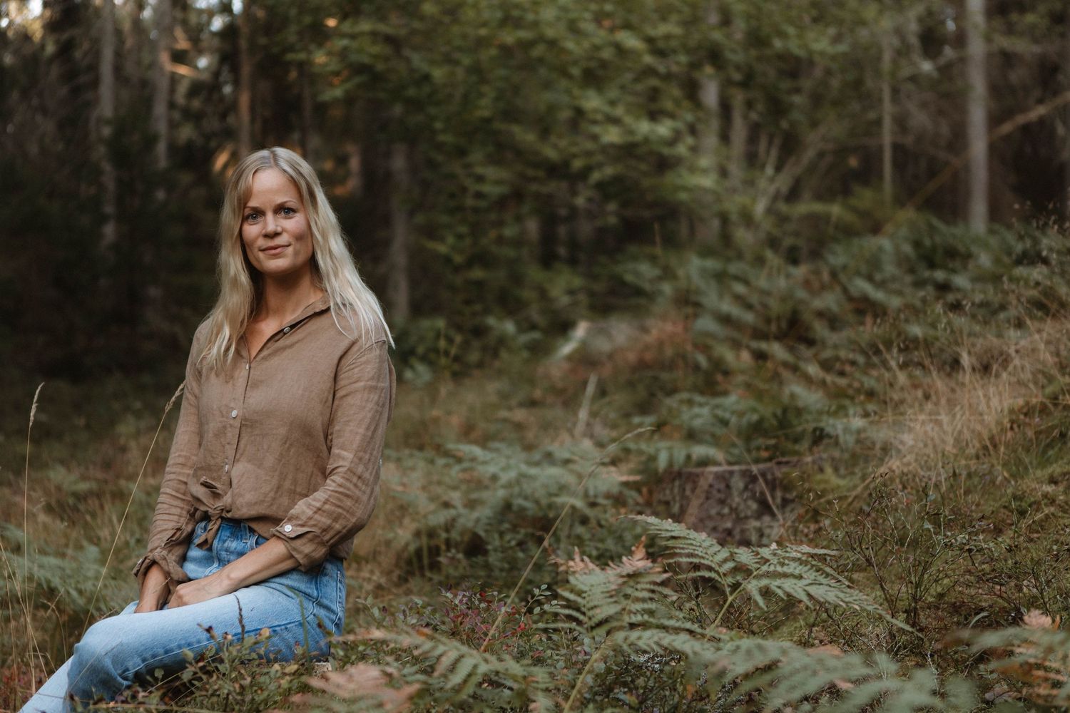 Person in casual attire sits thoughtfully among ferns in forest setting.