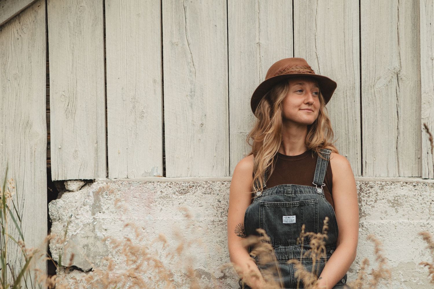Casual countryside portrait against white wooden wall wearing overalls and brown hat.
