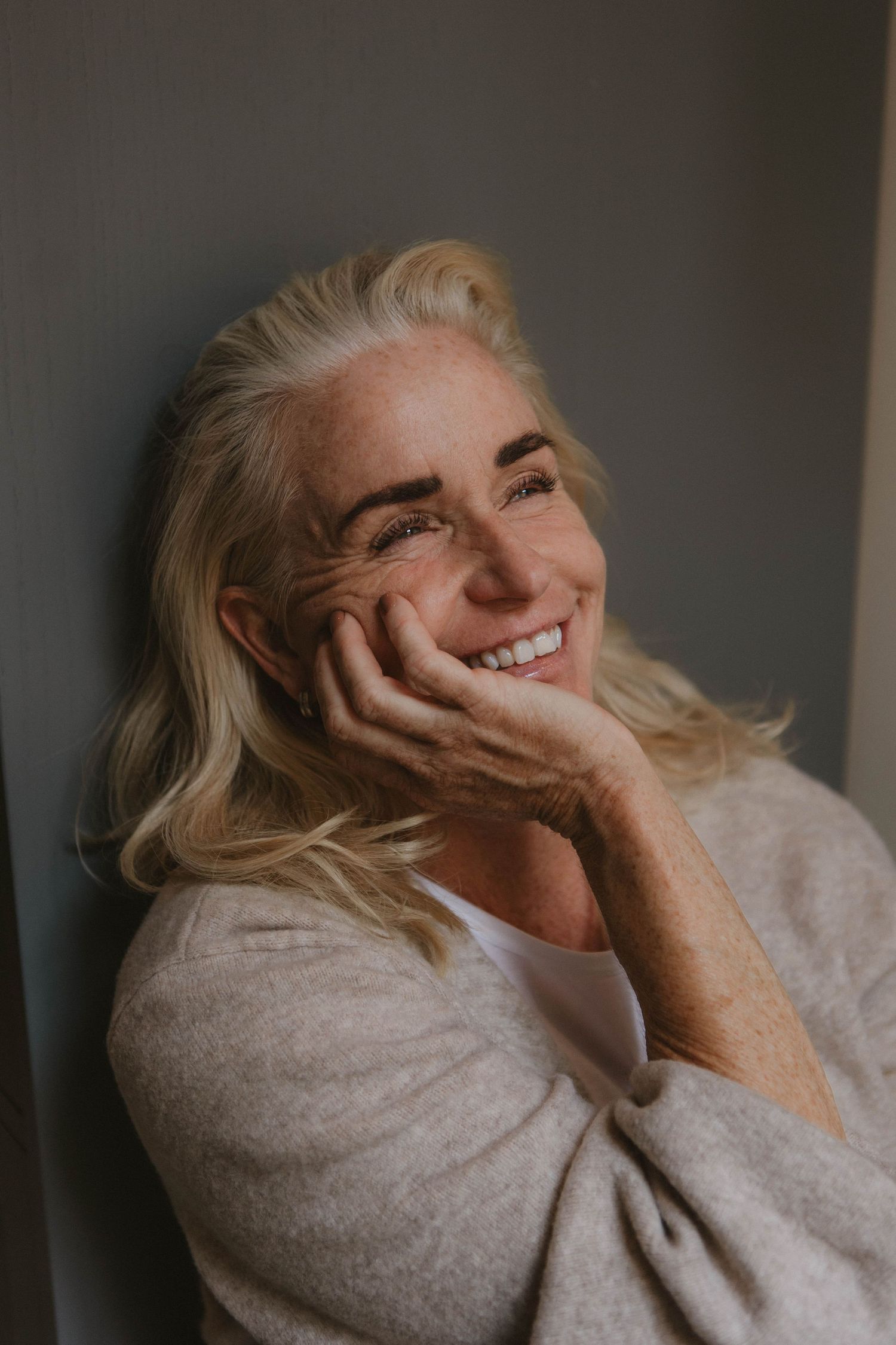 Portrait of someone with blonde hair wearing a gray sweater smiling warmly while resting their face on their hand.