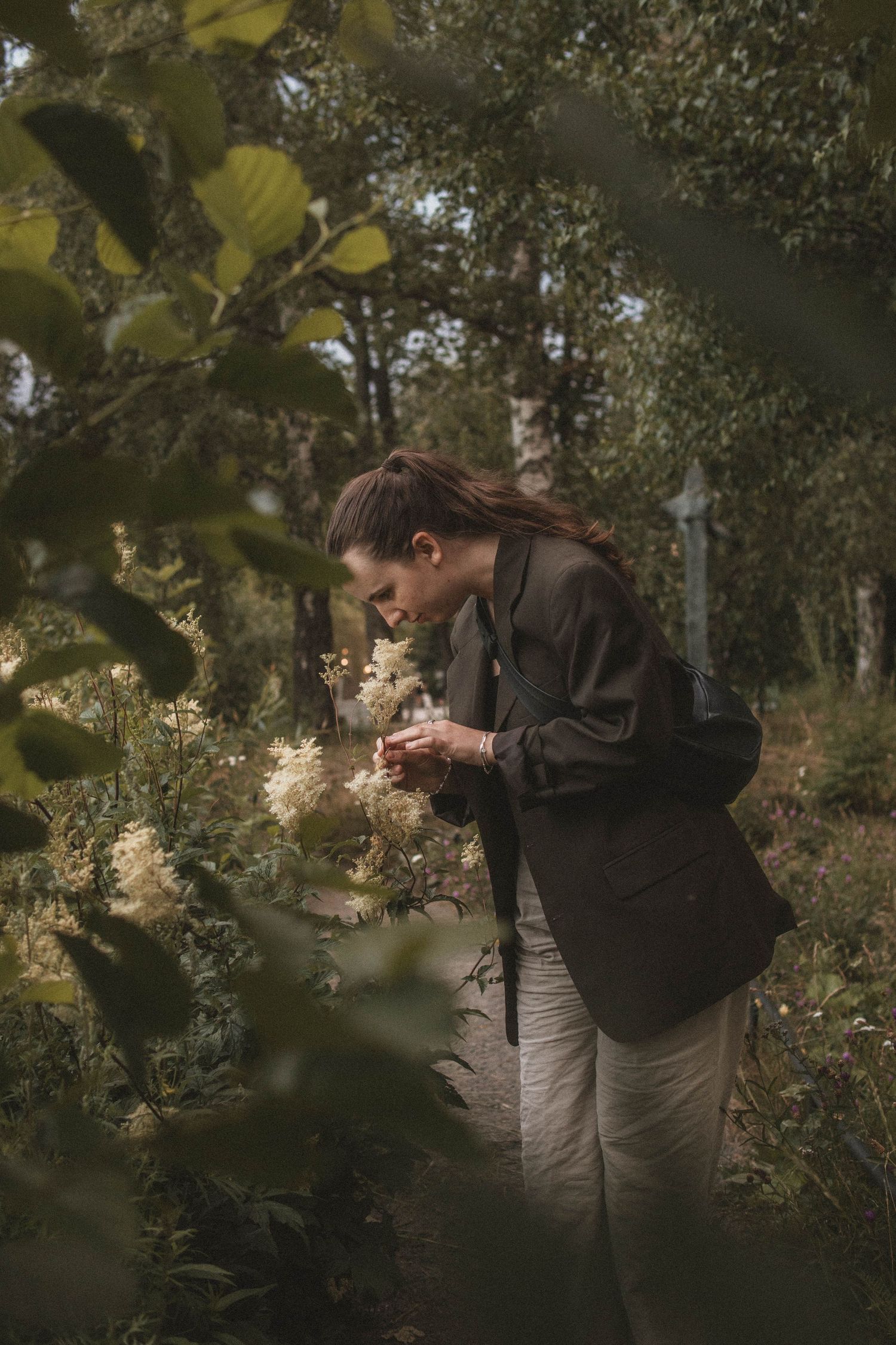 A person in a brown jacket bends down to smell delicate white wildflowers in a dreamy forest setting.