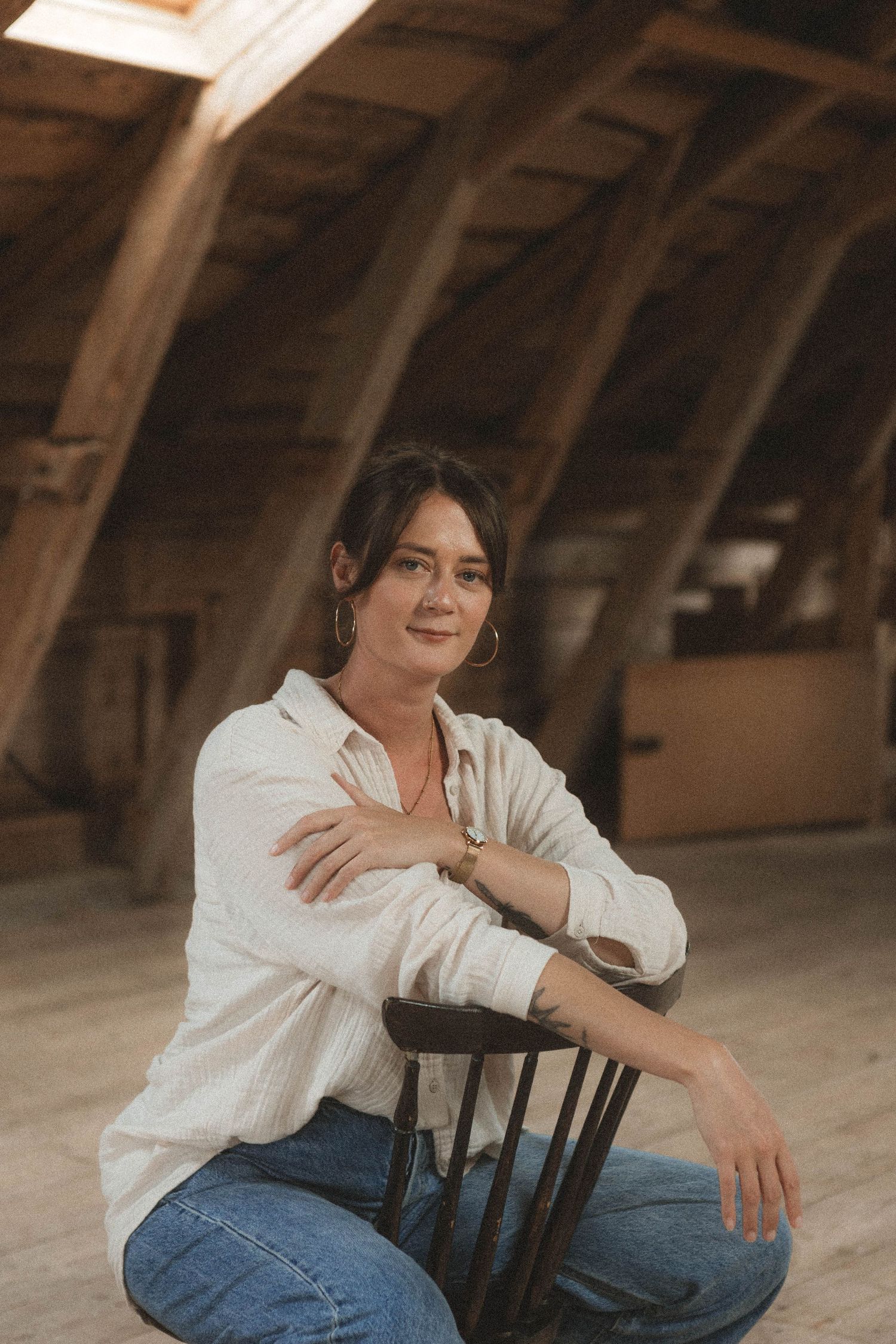Figure lounging casually in black wooden chair wearing white button-down shirt in wooden attic setting.