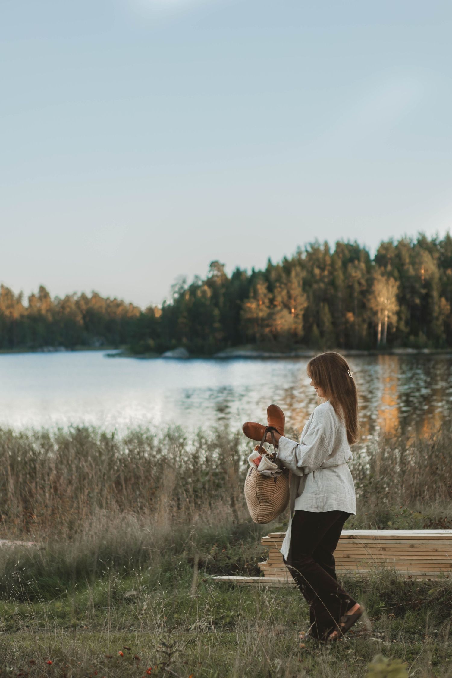 Figure in white stands by lakeside with a basket among tall grasses during golden hour.