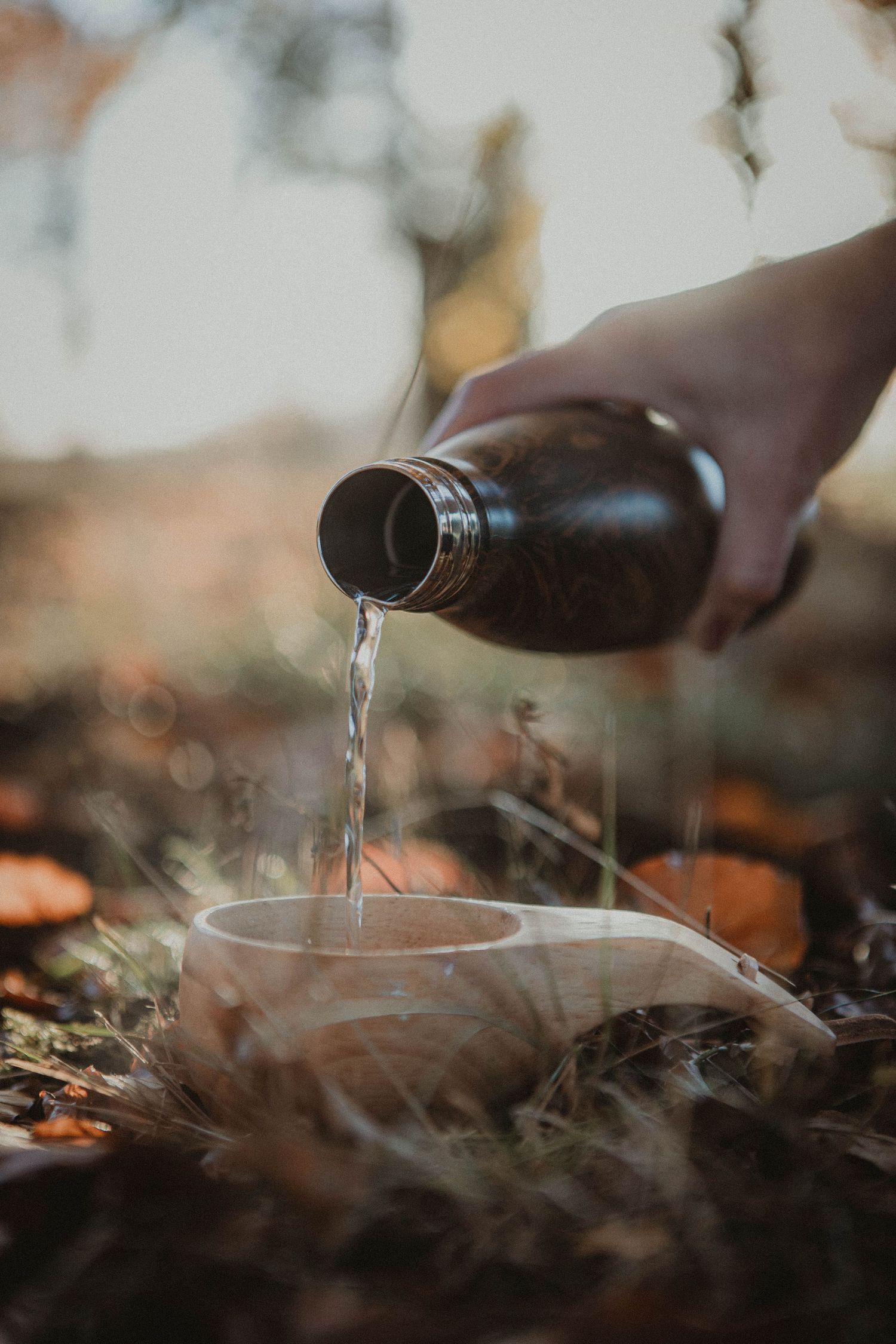 A hand pours steaming liquid from a dark bottle into a misty cup surrounded by autumn leaves in soft natural light.