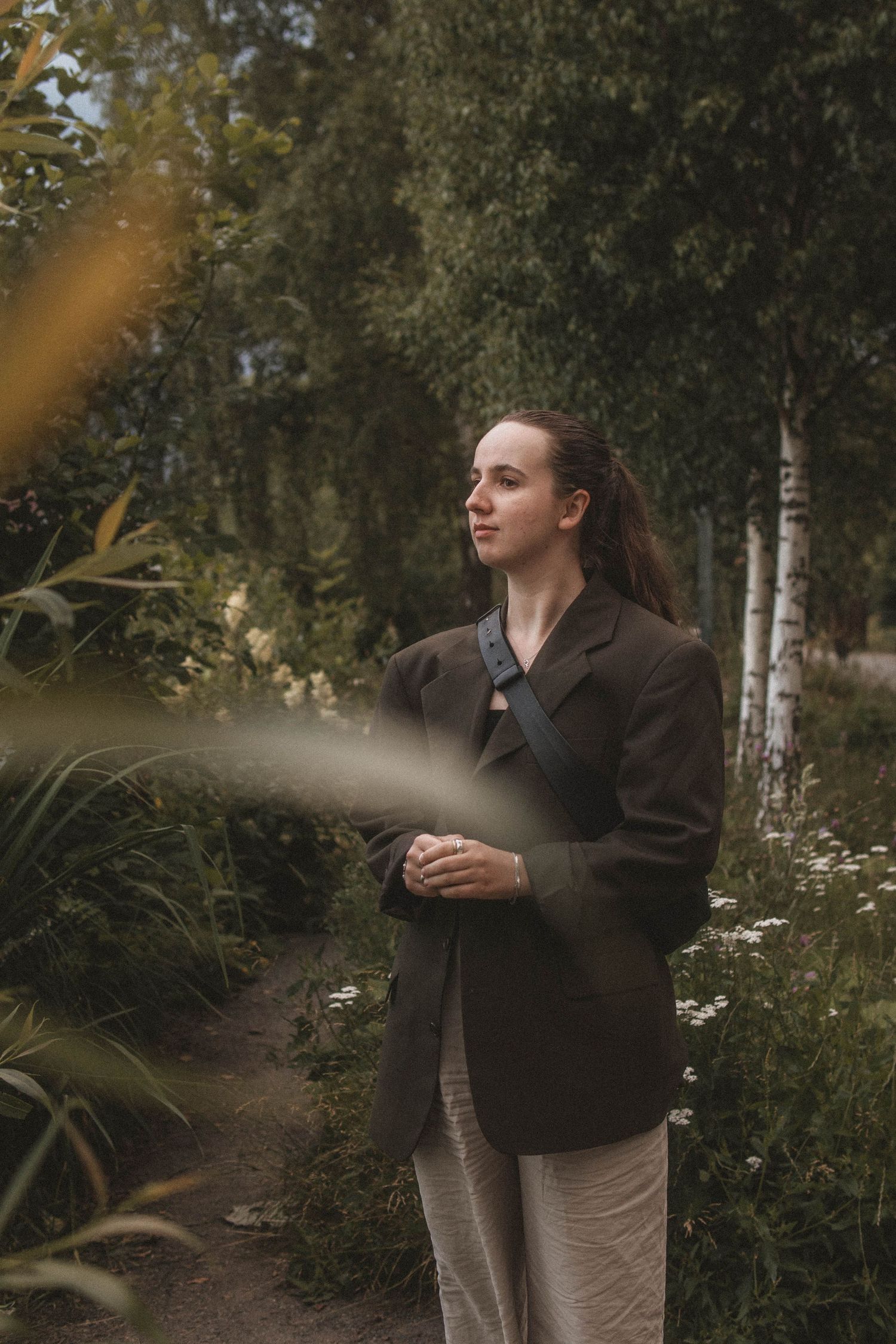 A person in a dark blazer stands thoughtfully in a wooded area with birch trees and wildflowers.