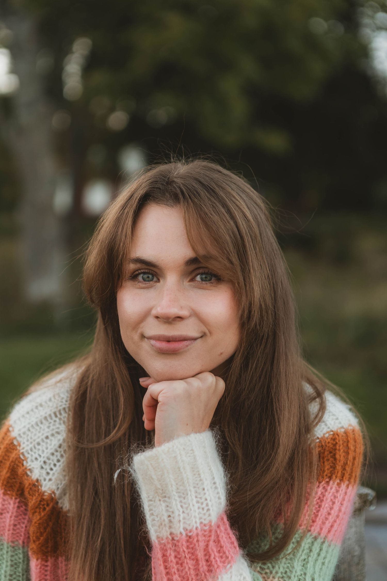 A person wearing a colorful striped knit sweater poses outdoors with their chin resting on their hand.
